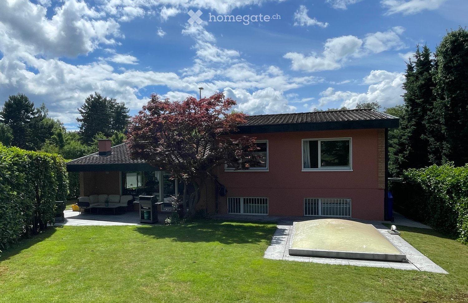 Single-story house with red brick exterior, surrounded by a well-maintained lawn and trees. Features include a patio area with outdoor furniture and a barbecue, and a small raised platform.