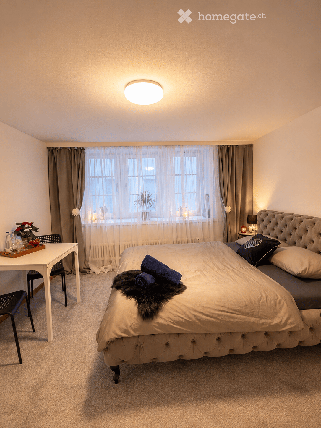 Cozy bedroom with a large bed, nightstand, table, and chair. The room features a large window with curtains, a round ceiling light, and a decorative tray on the table.