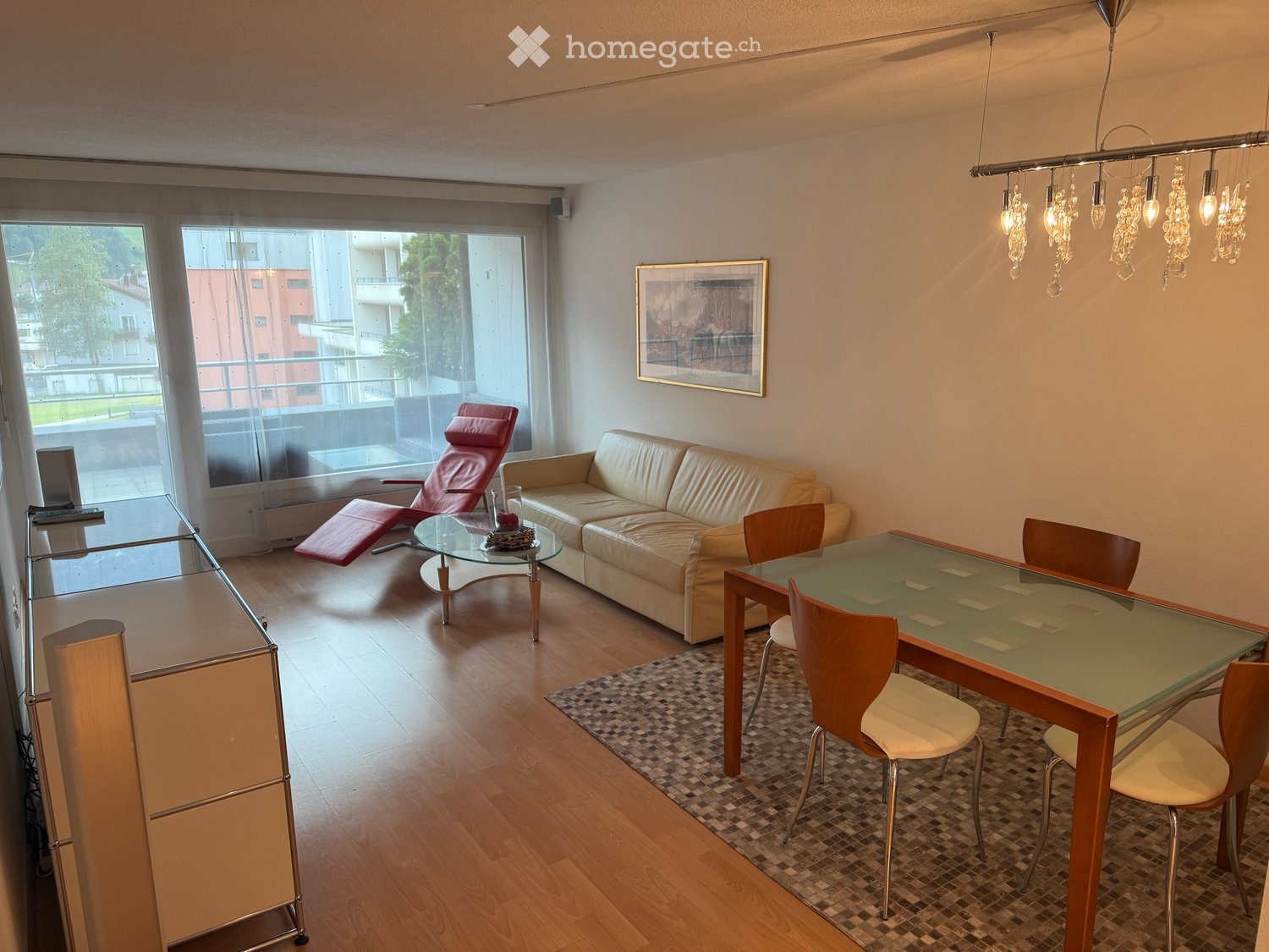 Modern living room with a cream leather sofa, red leather chair, glass coffee table, dining table with chairs, and a hanging light fixture.