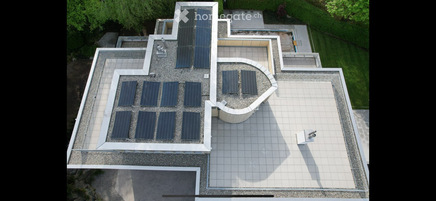 Aerial view of a modern building roof with multiple solar panels arranged in a grid pattern, surrounded by a gravel surface and a small garden area.
