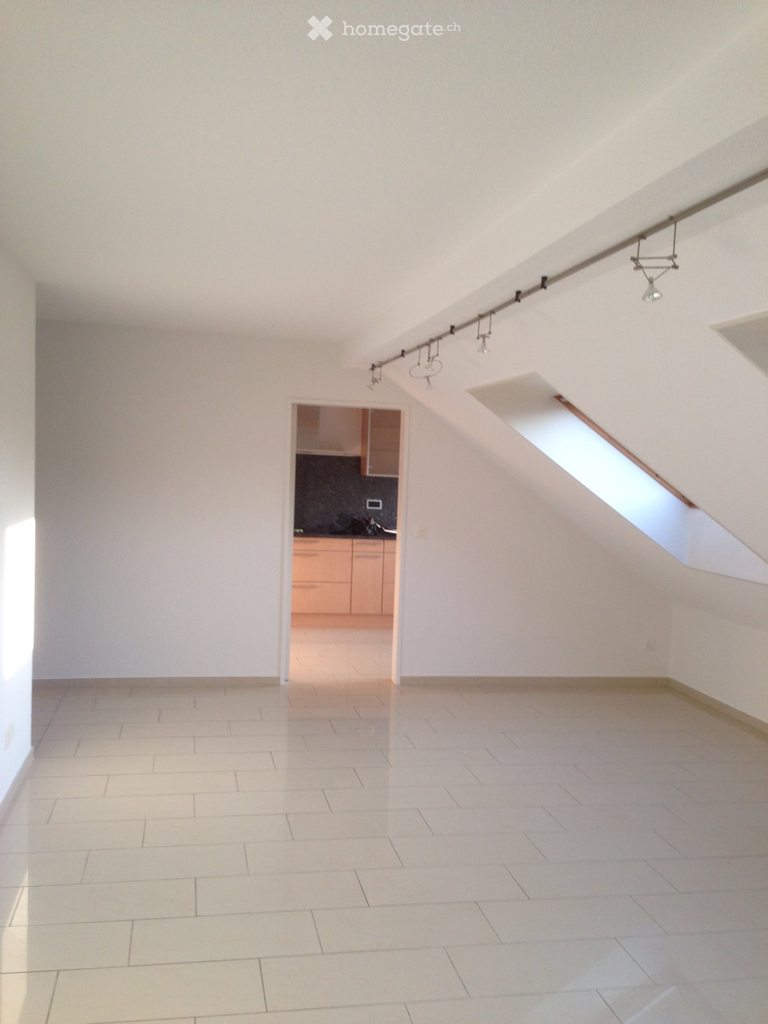 Empty room with white walls, tiled floor, and a skylight window. A small kitchen area is visible in the background with cabinets and a countertop.