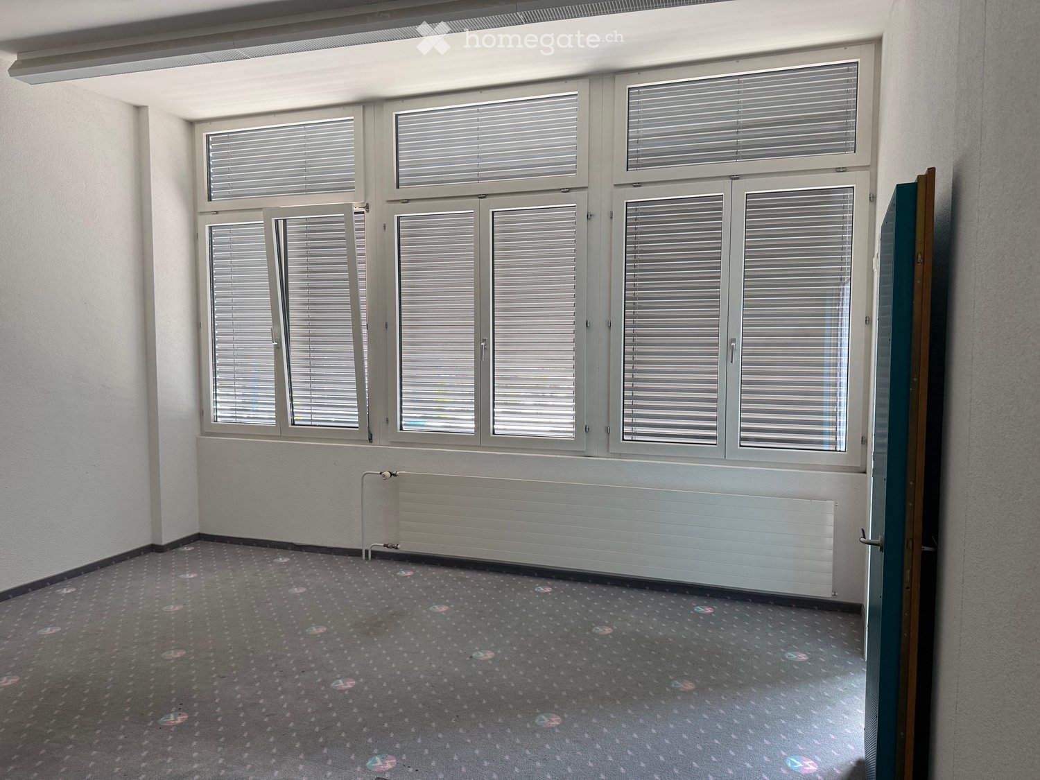 Empty room with grey carpet, white walls, large windows with blinds, and a radiator below the windows.