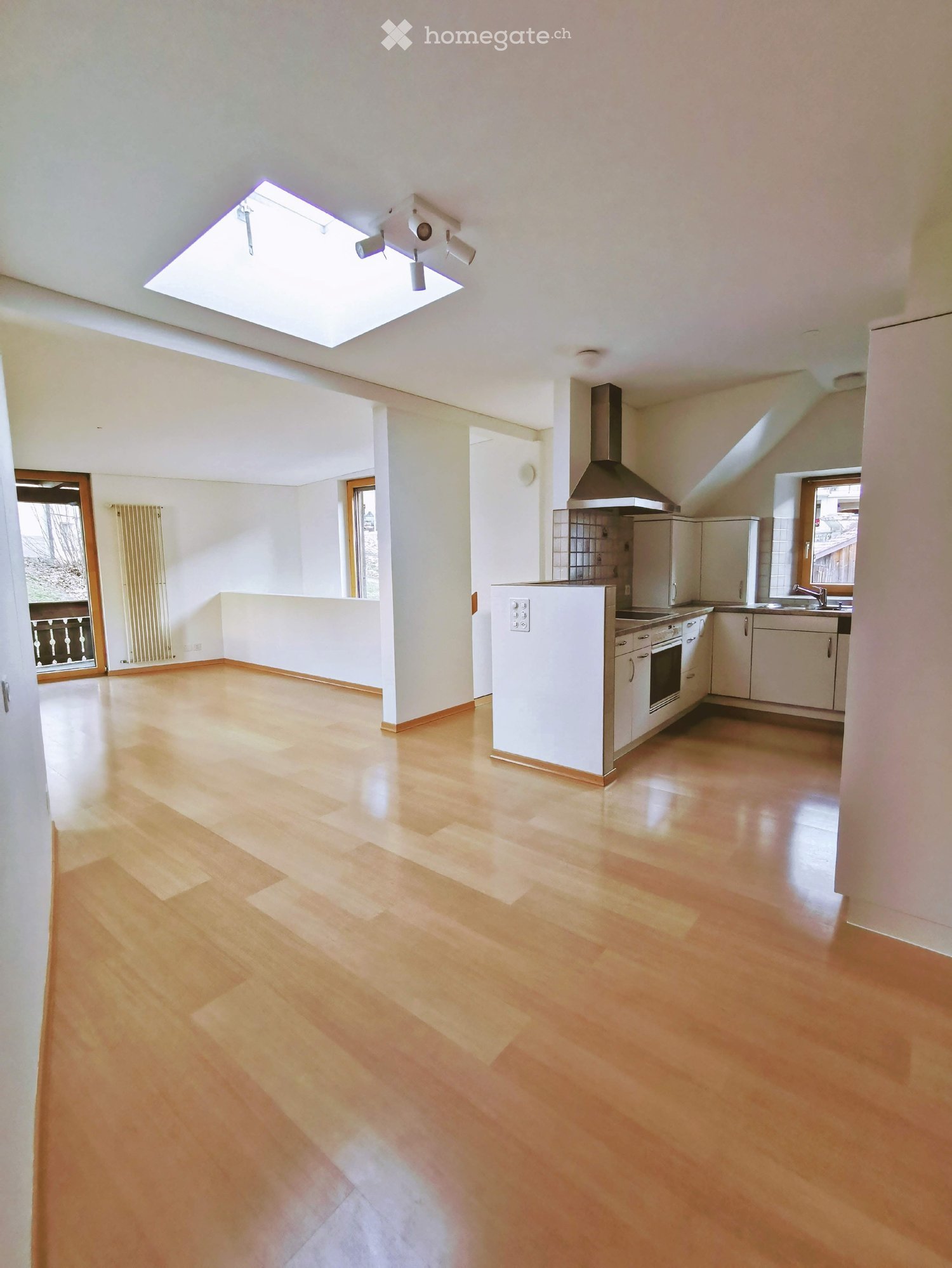 Open-plan living and kitchen area with light wooden flooring, white kitchen cabinets, stainless steel appliances, and large windows allowing natural light.