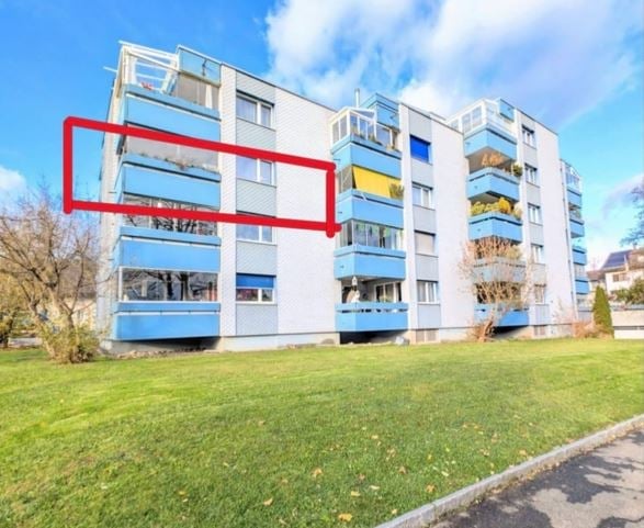 Multi-story apartment building with blue balconies and white walls, surrounded by green lawn.