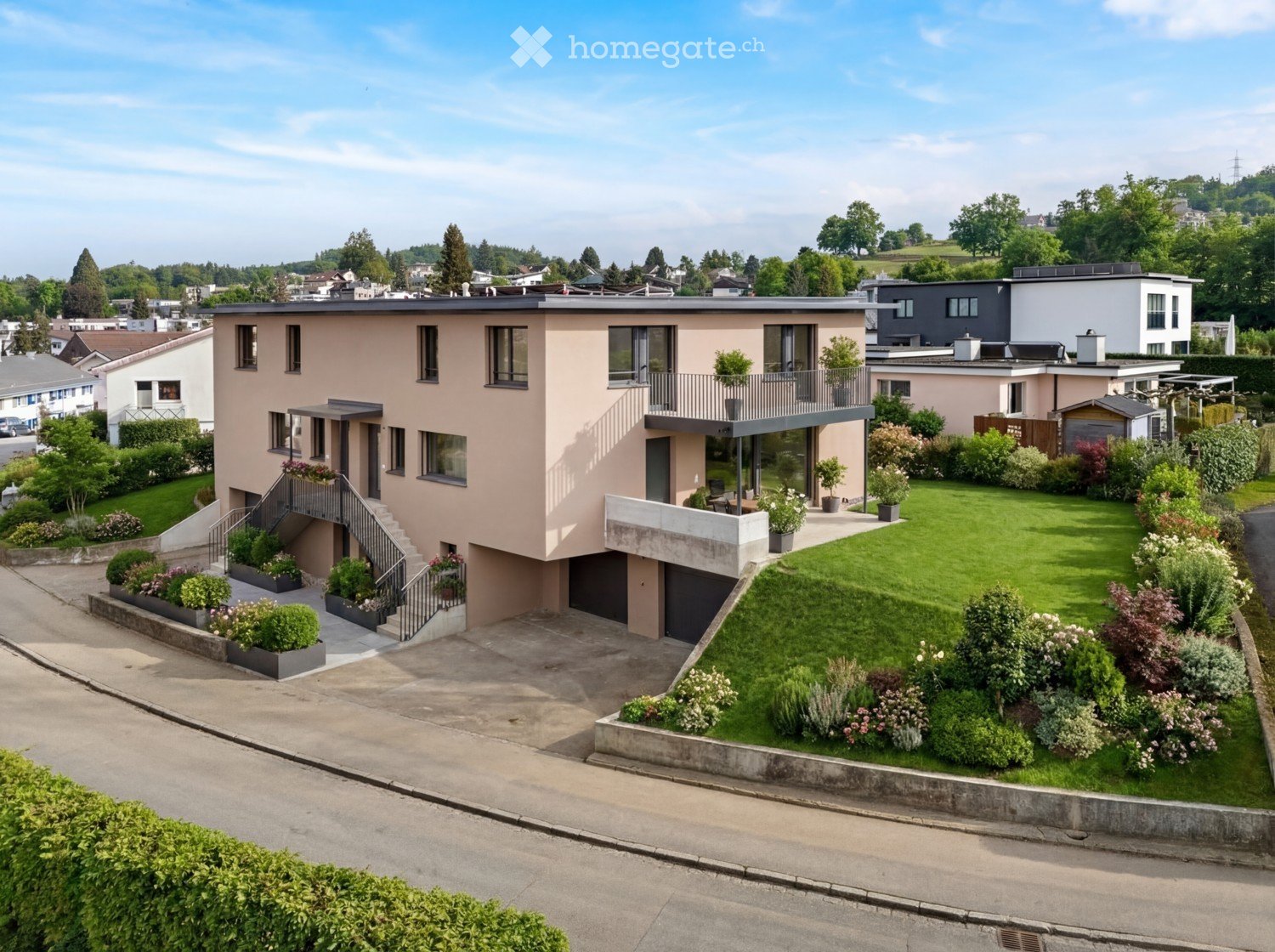 Modern two-story house with a raised ground floor, garage, and balcony. The house features a light pink facade, large windows, and a well-maintained garden with various plants and shrubs.