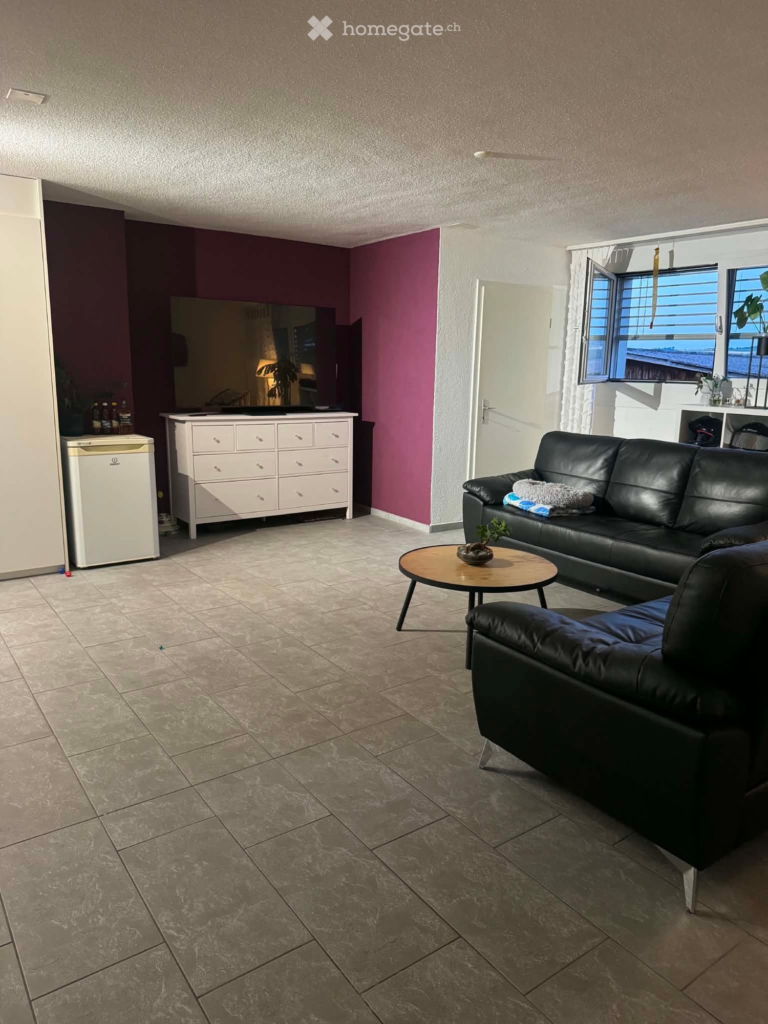 Living room with a black leather sofa, a round wooden coffee table, a dresser, and a refrigerator. The room has tiled flooring and large windows with blinds.