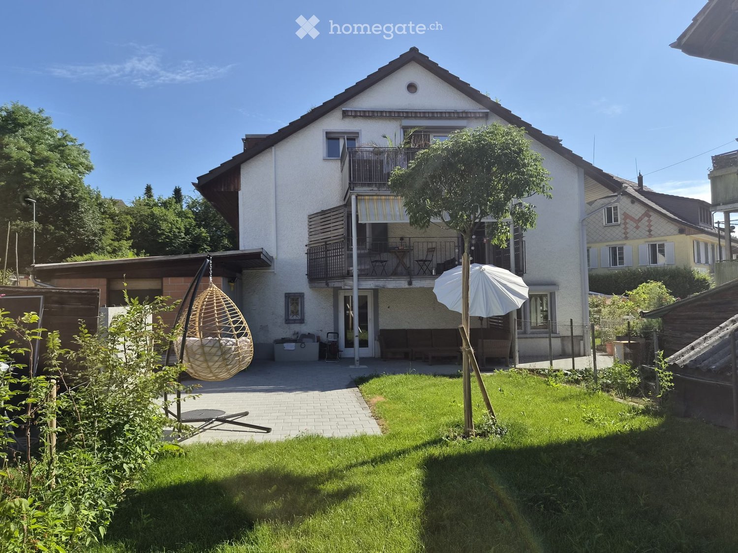 A two-story house with a balcony, surrounded by a well-maintained garden with a hanging chair, umbrella, and outdoor seating area.
