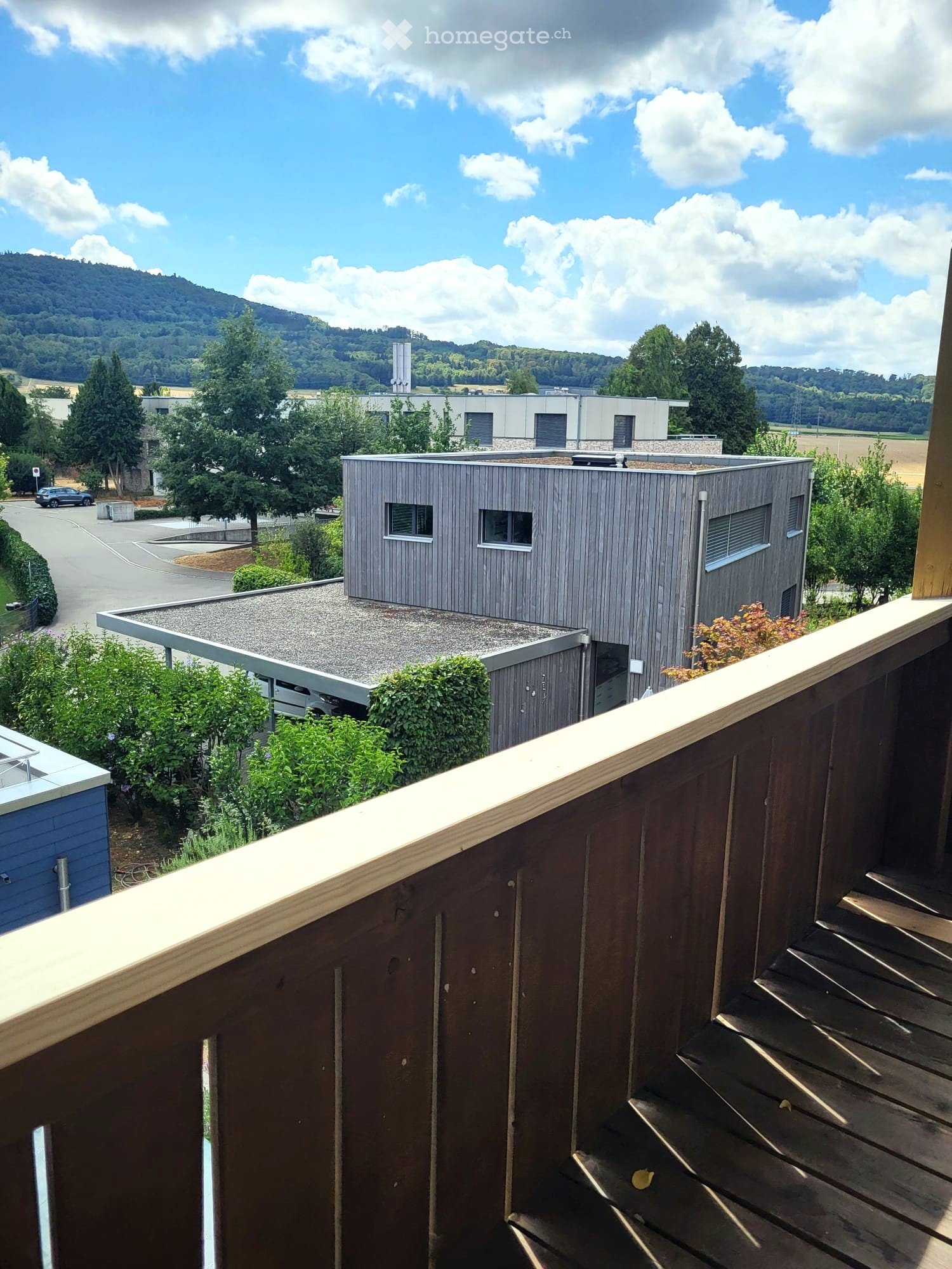 Modern building with grey wooden facade, flat roof, and small windows, surrounded by greenery and trees with a scenic hill in the background.