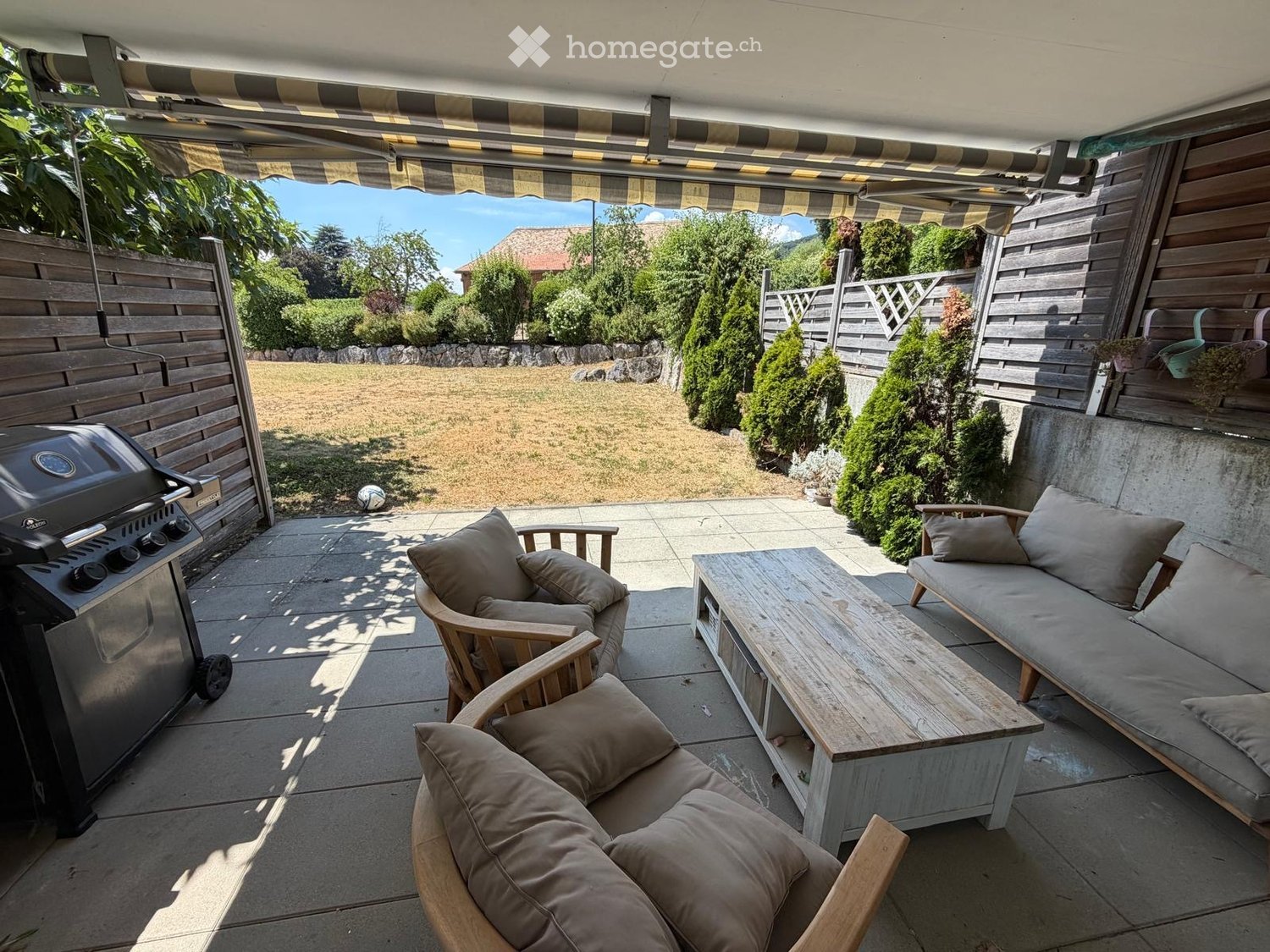 Outdoor patio with a covered seating area, featuring a sofa, chairs, a coffee table, and a grill, overlooking a grassy yard with trees and a fence.