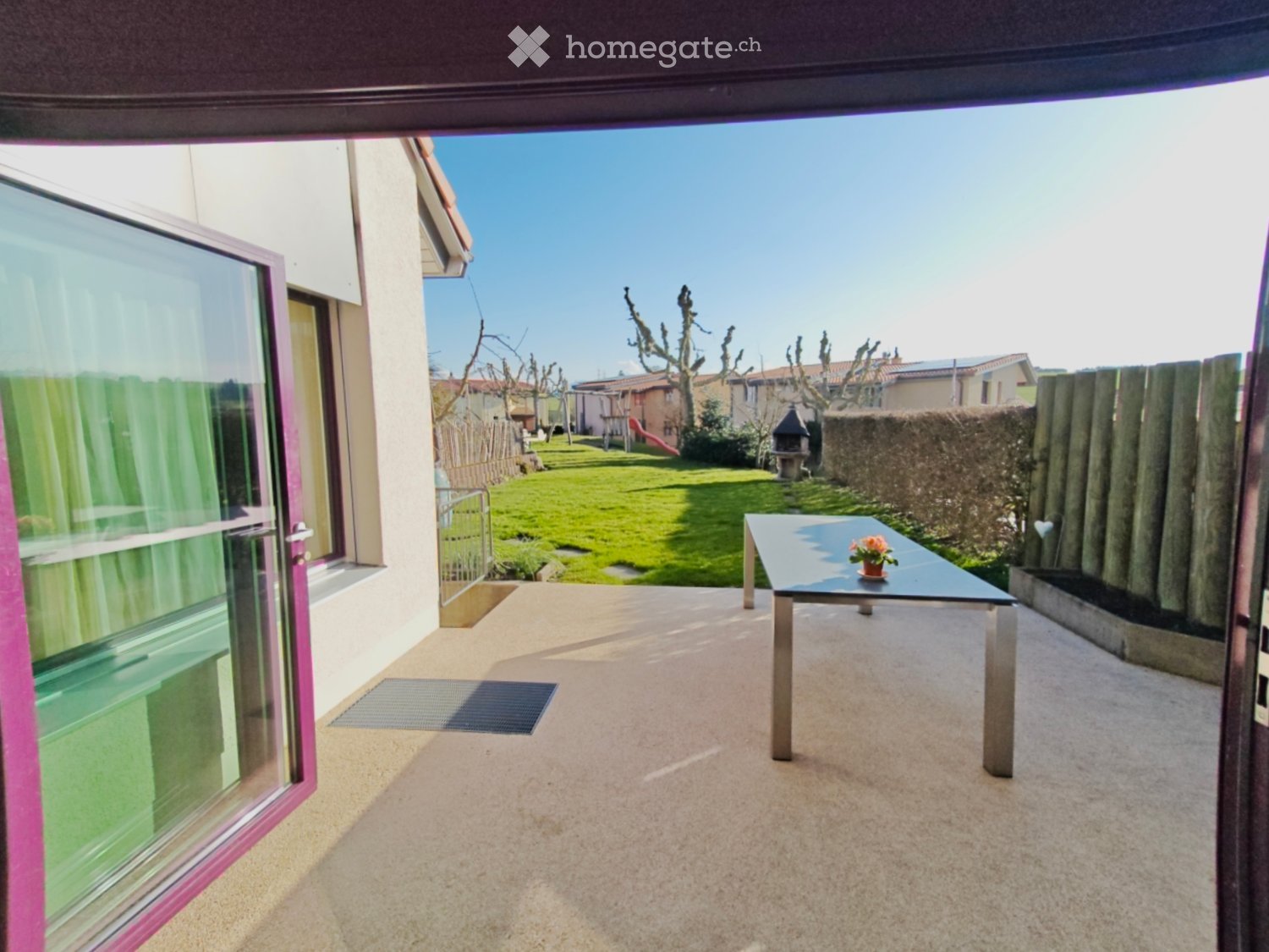 Balcony with a small table and flower pot, overlooking a grassy backyard with a wooden fence and neighboring houses.