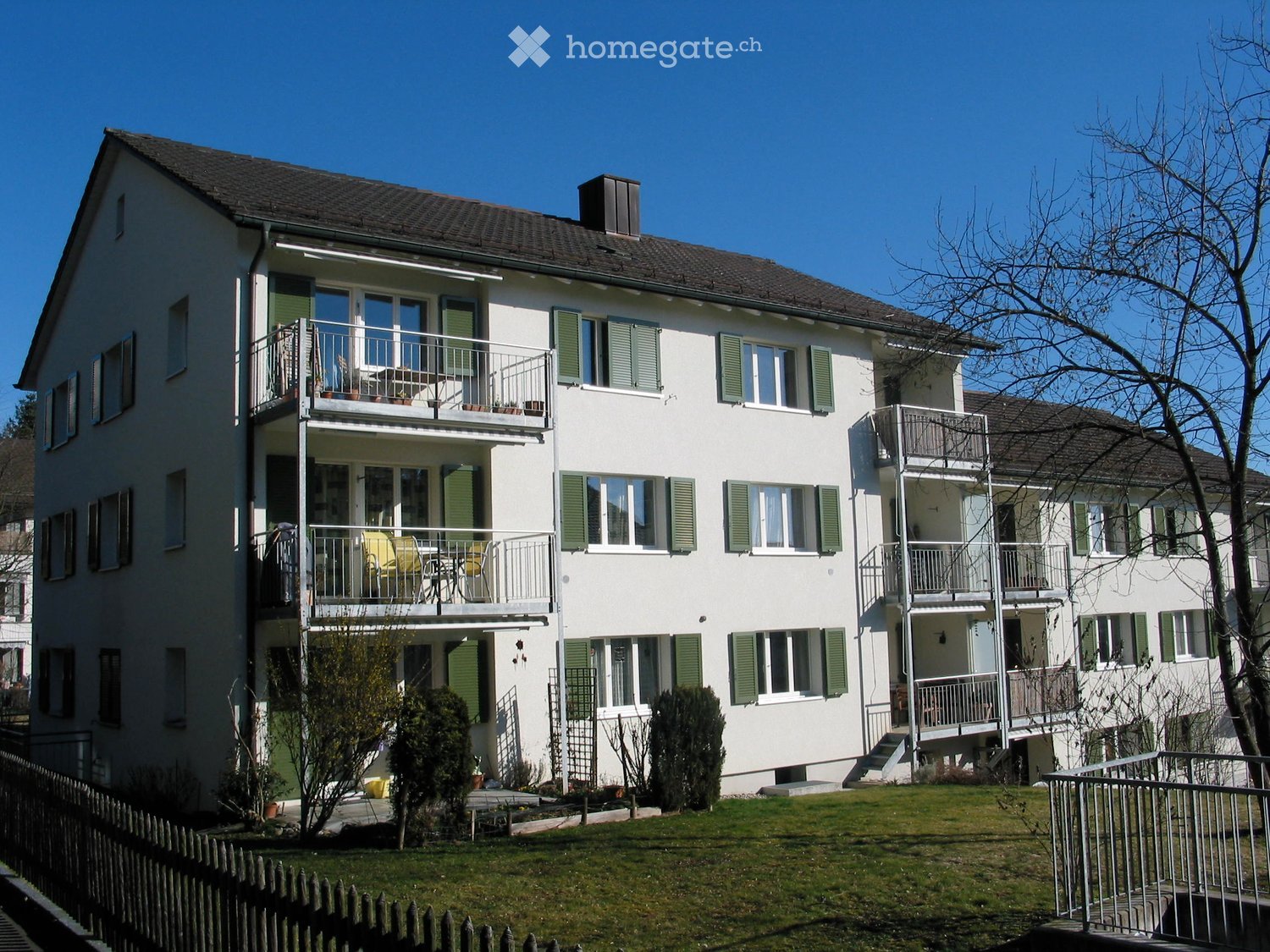 A two-story white house with green shutters, multiple balconies, and a well-maintained lawn.