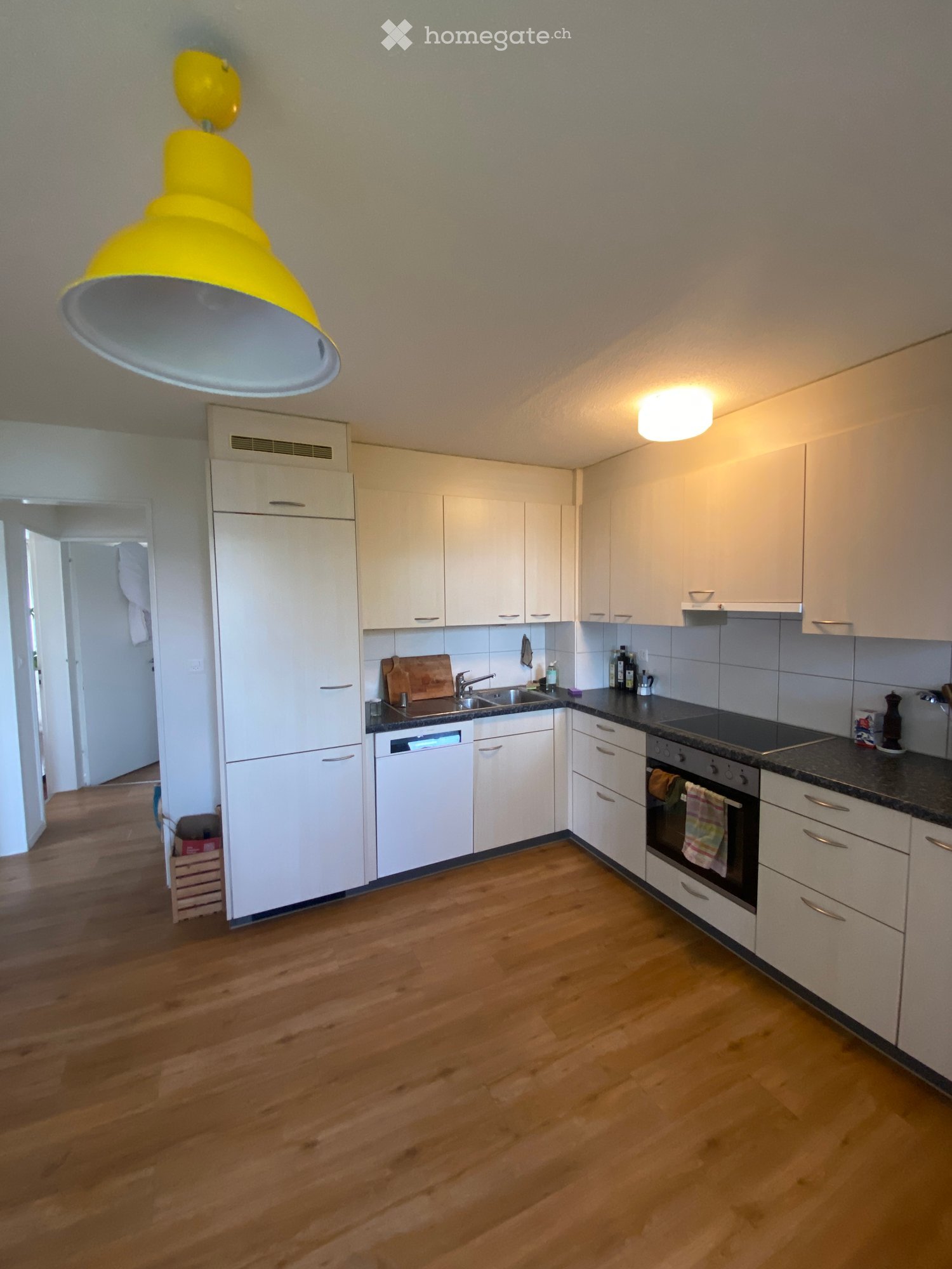 Modern kitchen with white cabinets, wooden flooring, and stainless steel appliances including a refrigerator, dishwasher, and oven. A large yellow pendant light hangs from the ceiling.
