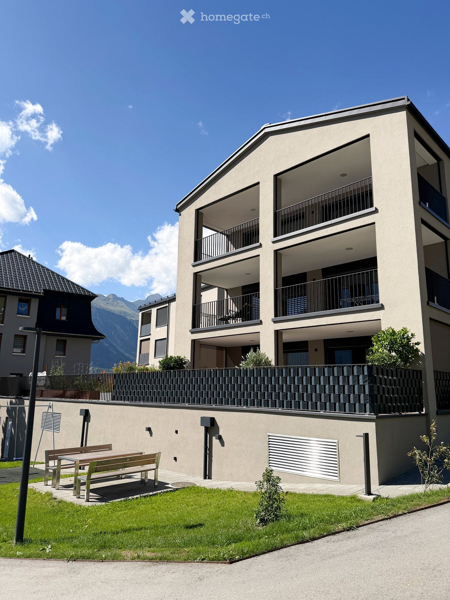 Modern two-story building with balconies, surrounded by a small garden and a view of mountains.