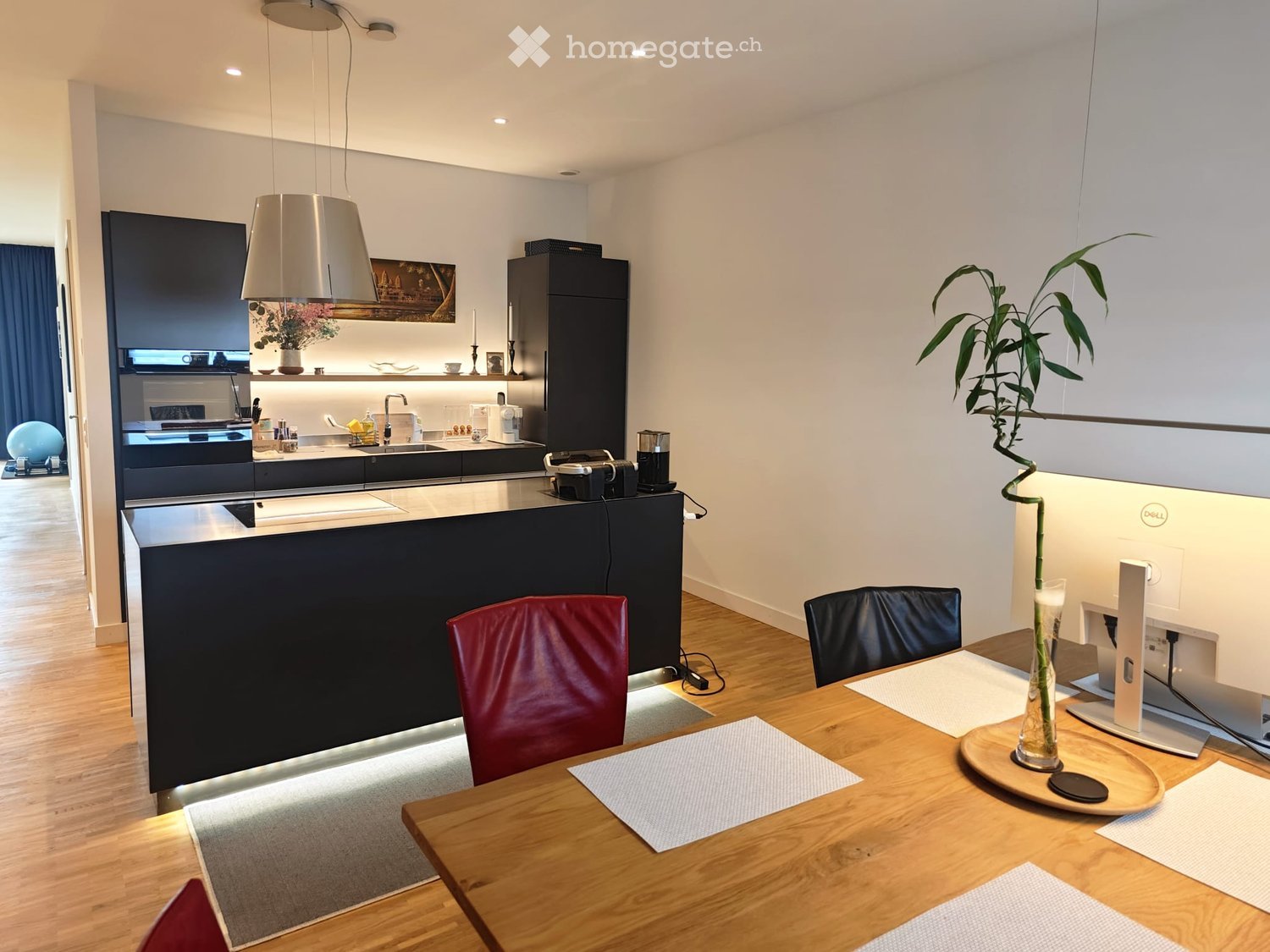 Modern kitchen and dining area with a wooden dining table, red chairs, a large screen, and a sleek black kitchen counter with built-in appliances.