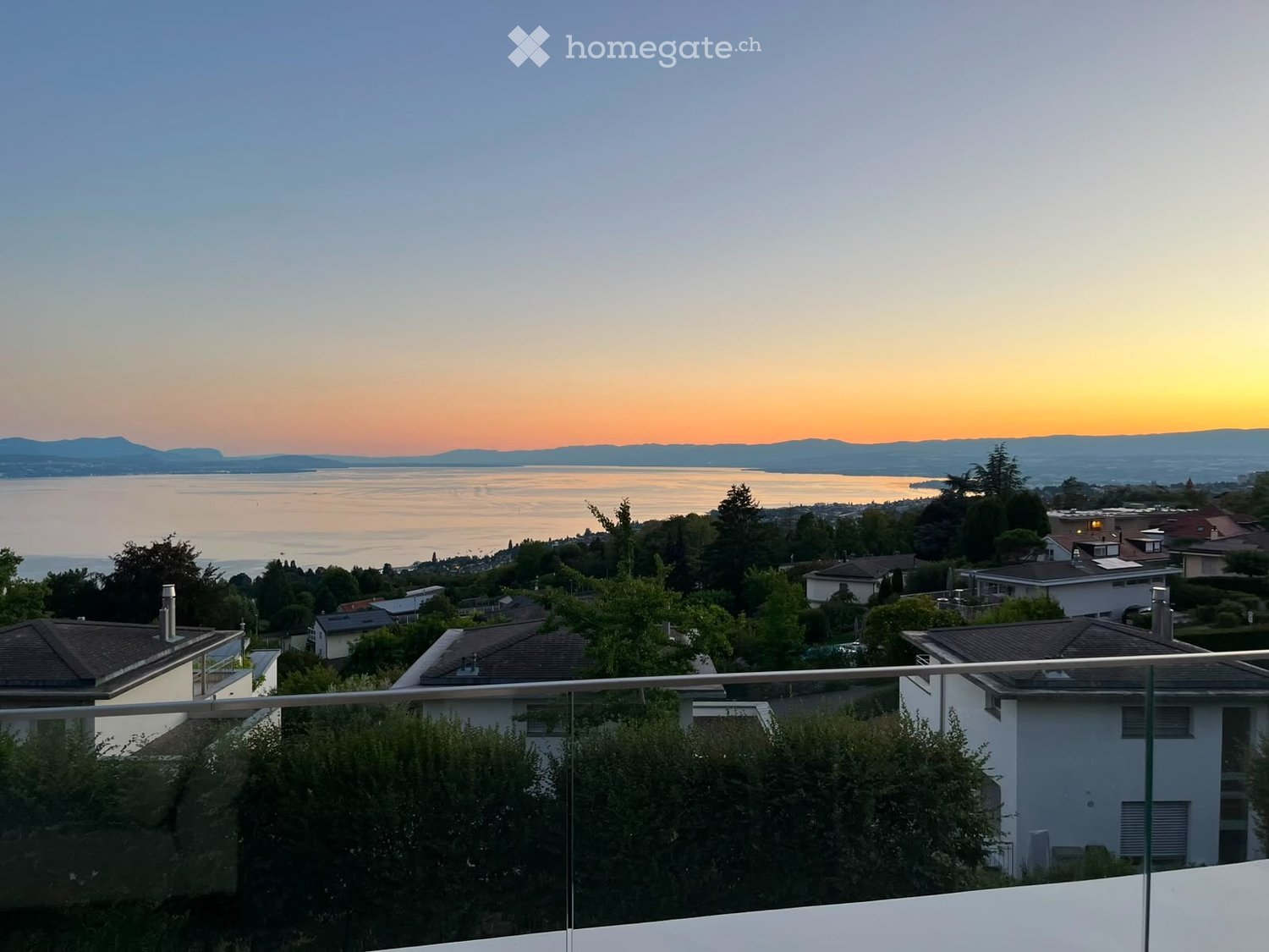 Balcony overlooking a scenic view of a lake and distant mountains during sunset, with surrounding houses and greenery.