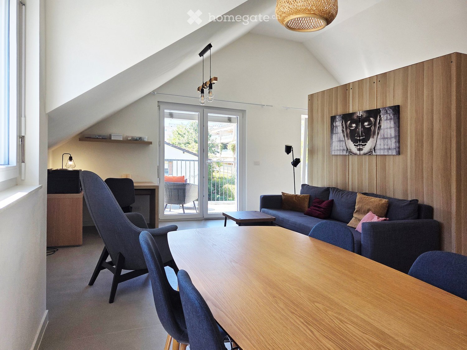 Modern living room with a slanted ceiling, large windows, and a sliding glass door leading to a balcony. Features a blue sofa, wooden coffee table, dining table with chairs, and a decorative picture on the wall.