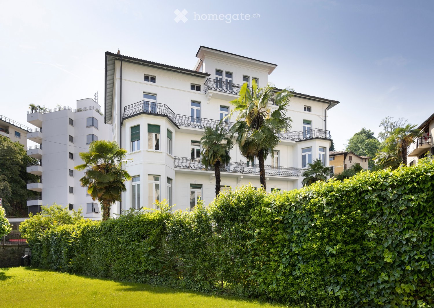 A multi-story white building with balconies and large windows, surrounded by lush greenery and palm trees.