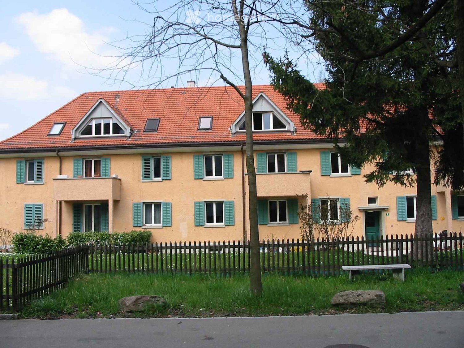 Two-story house with a red-tiled roof, light yellow walls, green shutters, and multiple windows. The house is surrounded by a wooden fence, trees, and a grassy yard.