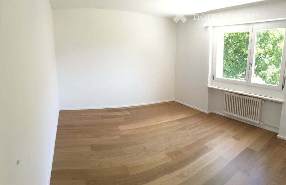 Empty room with light wooden flooring, white walls, and a large window with a view of greenery. A radiator is visible under the window.