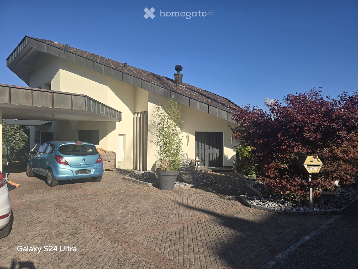 Two-story modern house with a flat roof, large windows, and a covered parking area. The driveway is paved with bricks, and there are potted plants near the entrance.