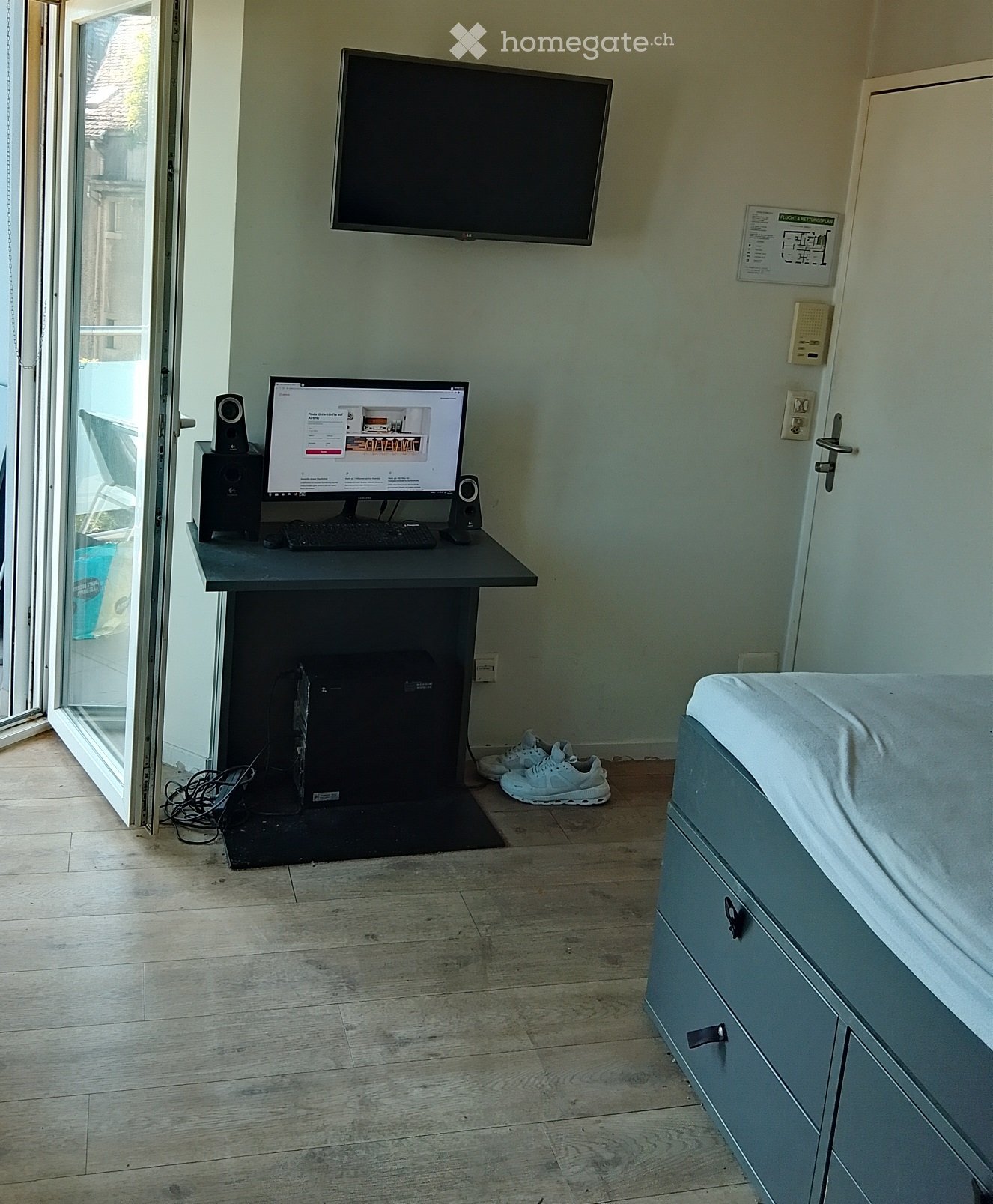 A furnished room with a desk setup, a television mounted on the wall, a drawer unit, and a pair of white sneakers on the floor.