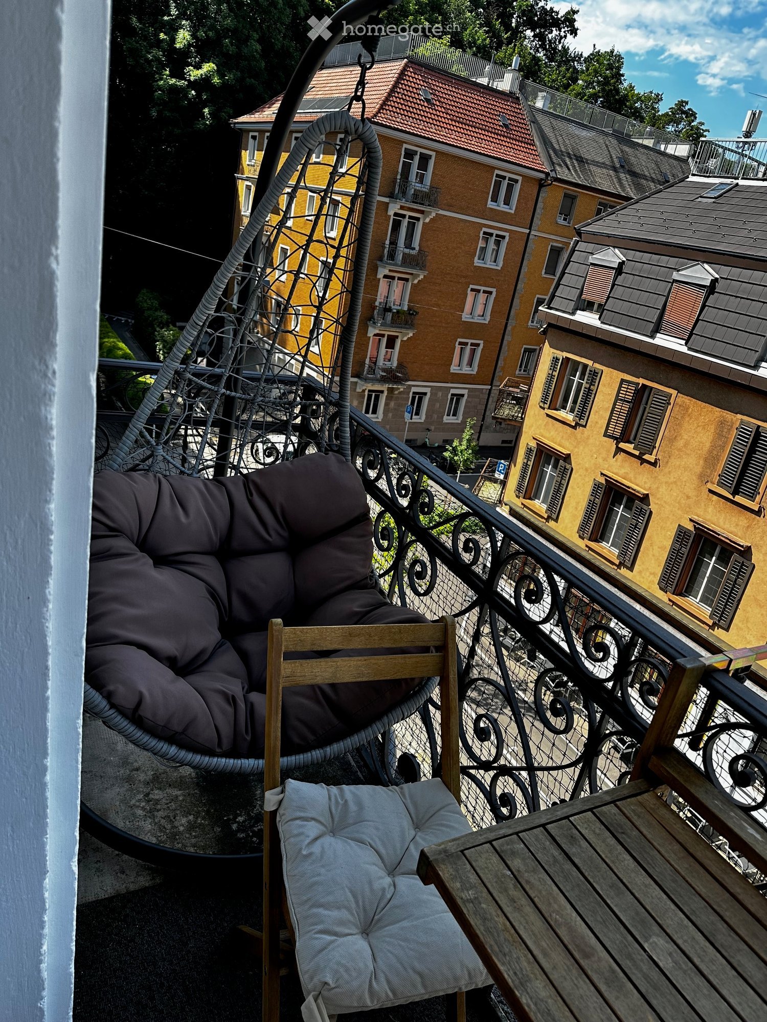 Balcony with a wooden table, a cushioned chair, and a hanging wicker chair, overlooking a cityscape with yellow buildings and balconies.