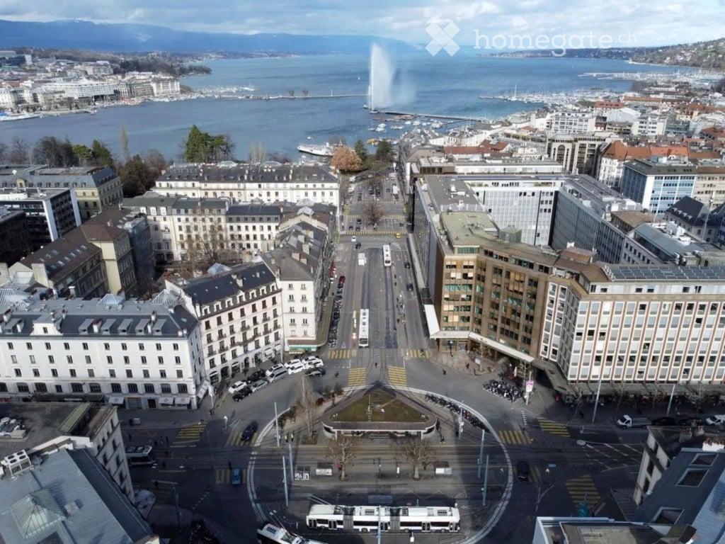 Aerial view of a cityscape with a prominent fountain in the distance, surrounded by buildings, roads, and a waterfront with boats.