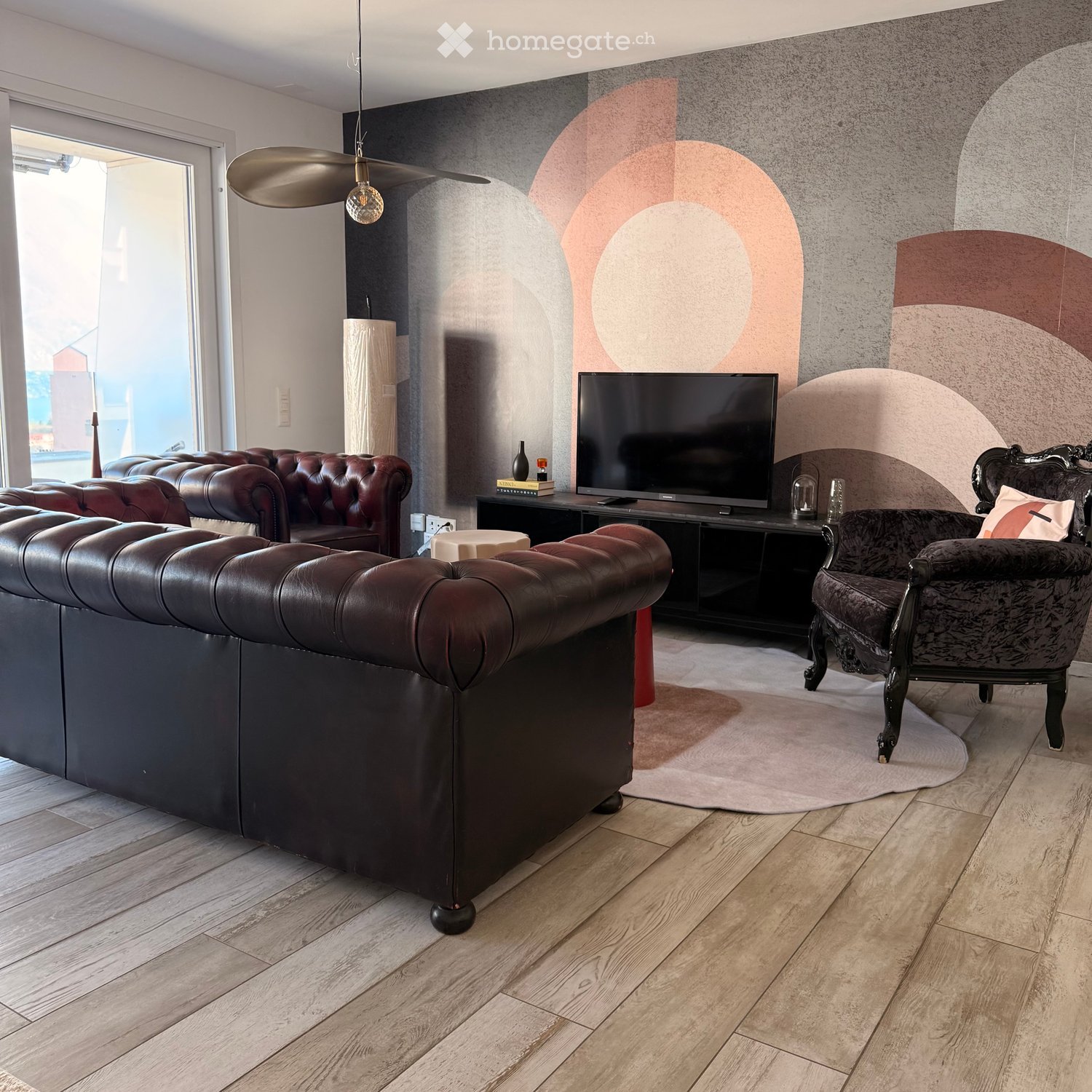 Modern living room with a large leather sofa, a patterned armchair, a TV stand with a flat-screen TV, a small round table, and a decorative lamp hanging from the ceiling.