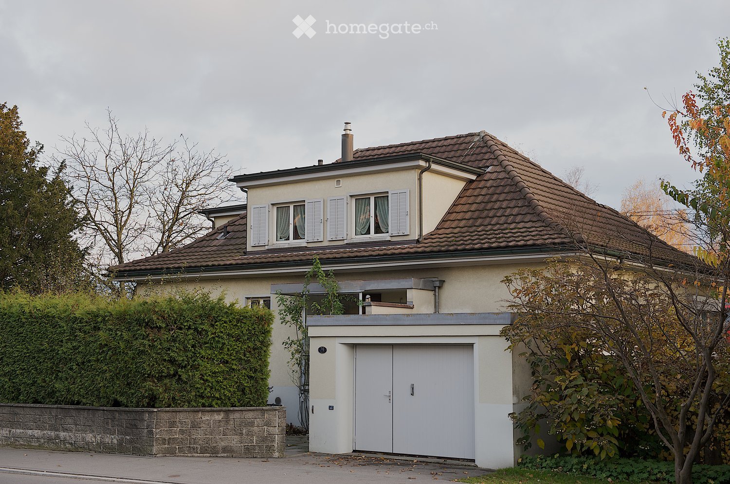Two-story house with beige walls, brown tiled roof, white garage door, and multiple windows with shutters.