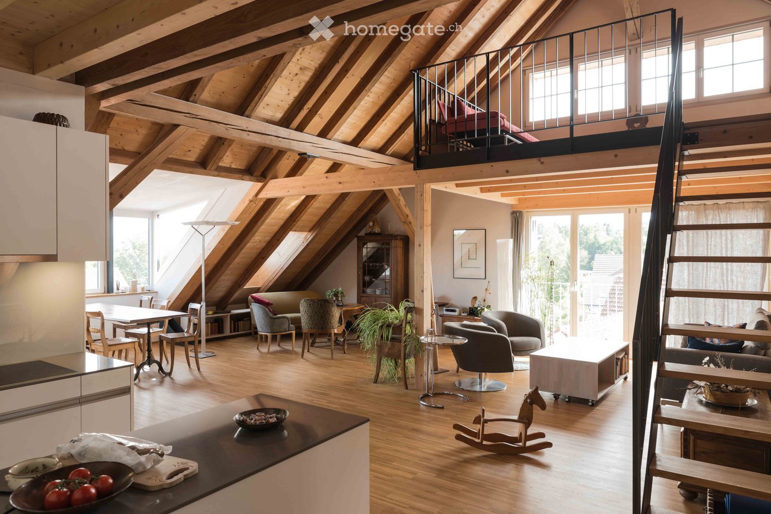 Spacious attic living area with wooden beams, exposed wooden ceiling, and large windows. Features a kitchen area with a counter, a dining table with chairs, a sofa, armchairs, a rocking horse, and a small balcony with a bed.