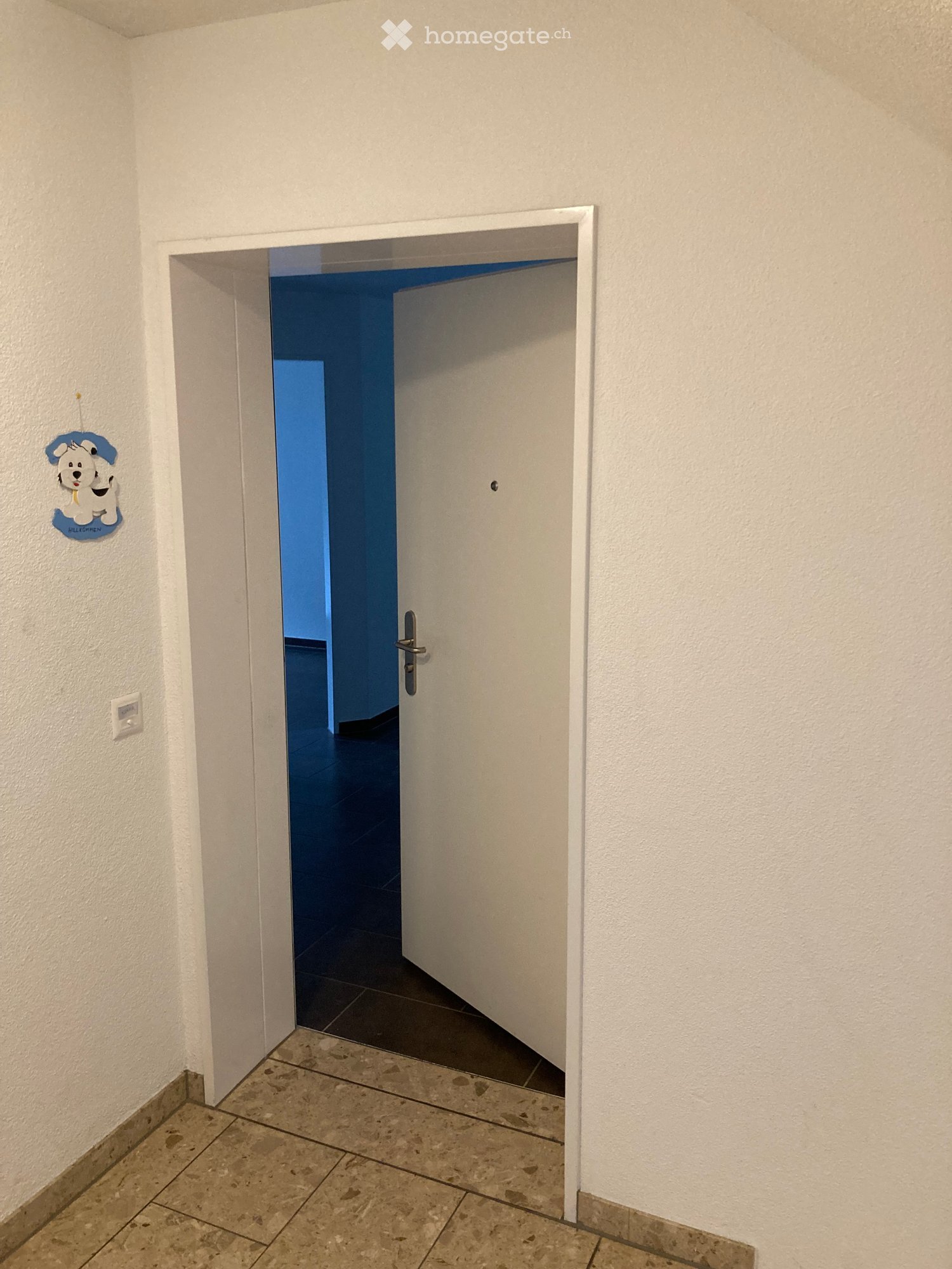 Empty hallway with white walls, tiled floor, and a partially open door leading to a blue-painted room.