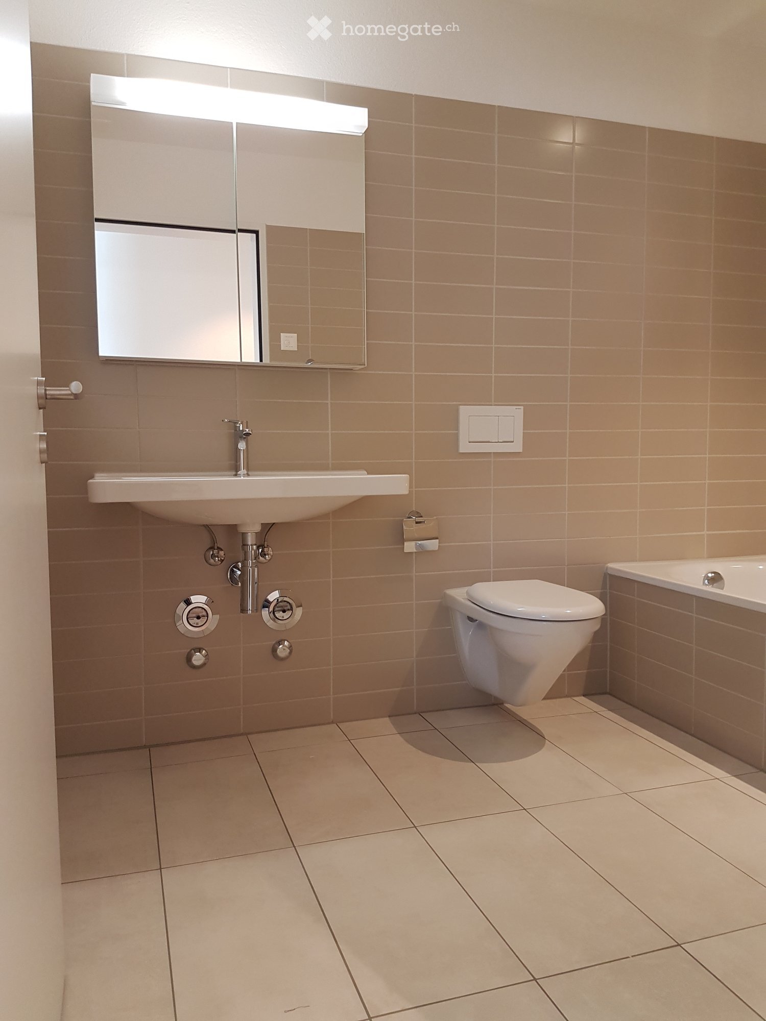 Modern bathroom with beige tiles, a white sink, a toilet, a bathtub, and a mirrored cabinet with built-in lighting.