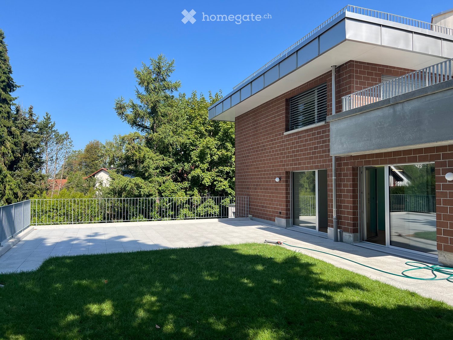 Modern two-story house with red brick facade, large windows, and a spacious balcony overlooking a well-maintained lawn and garden area.