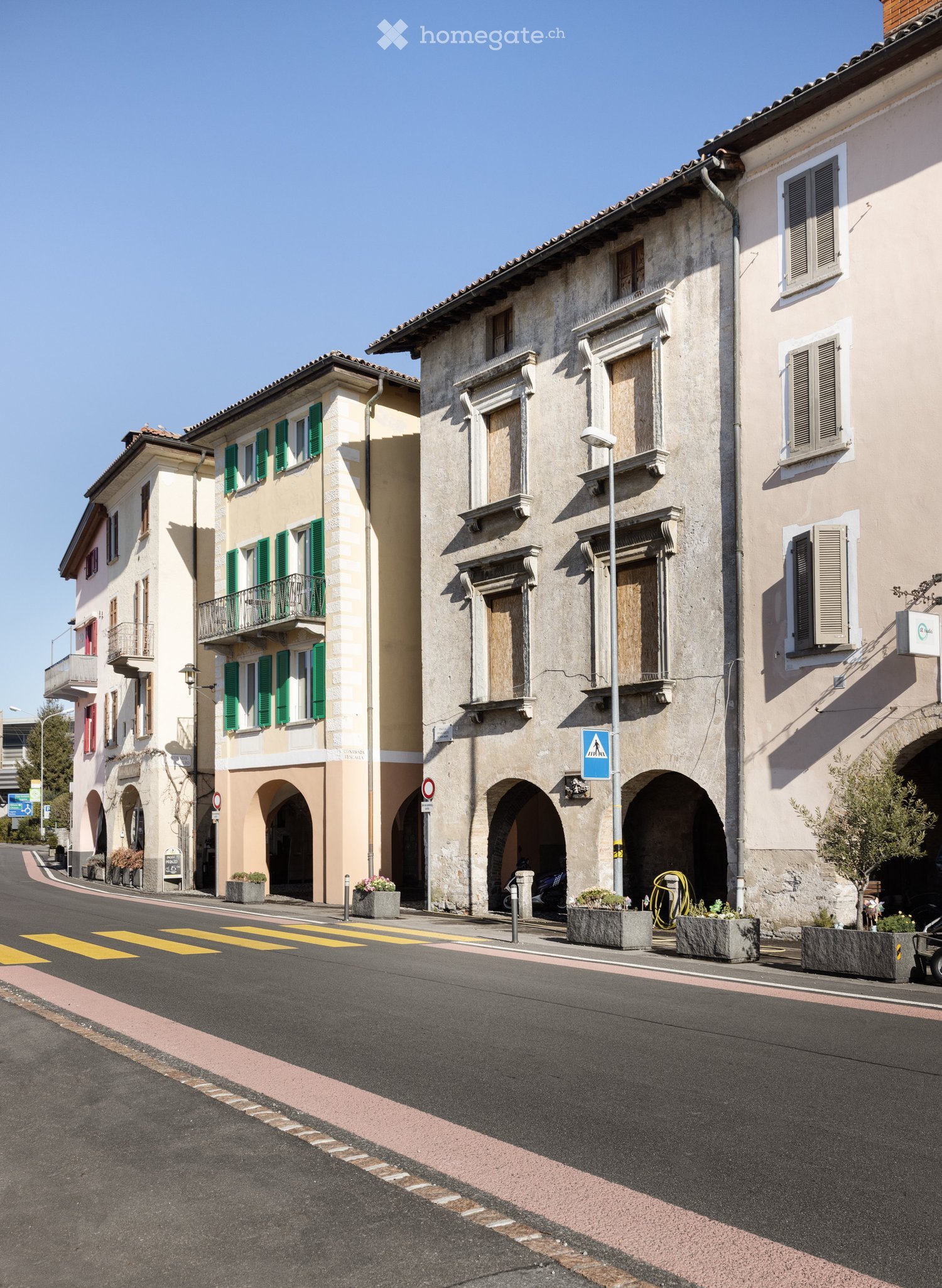 A street view of a European town with multi-story buildings, arched entrances, green shutters, and flower planters lining the sidewalk.