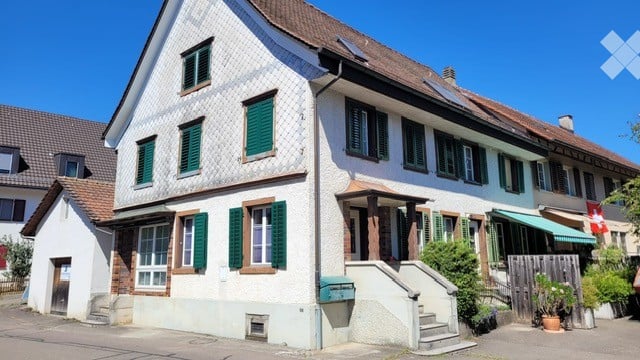 Traditional Swiss house with white walls, green shutters, and a red-tiled roof. The building has multiple windows and a small porch with steps leading up to the entrance.