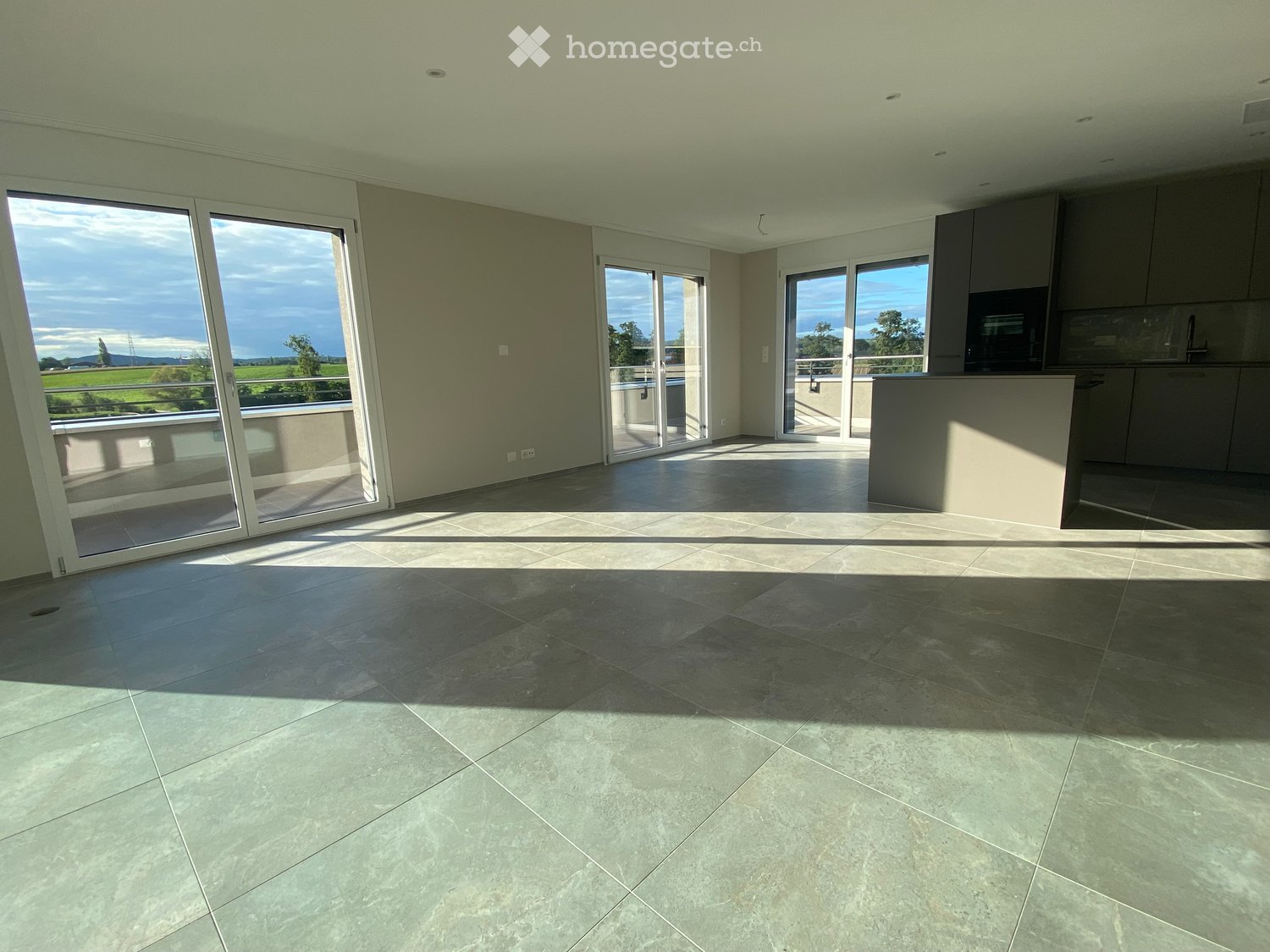 Empty room with large sliding glass doors leading to a balcony, tiled floor, and modern kitchen area visible in the background.