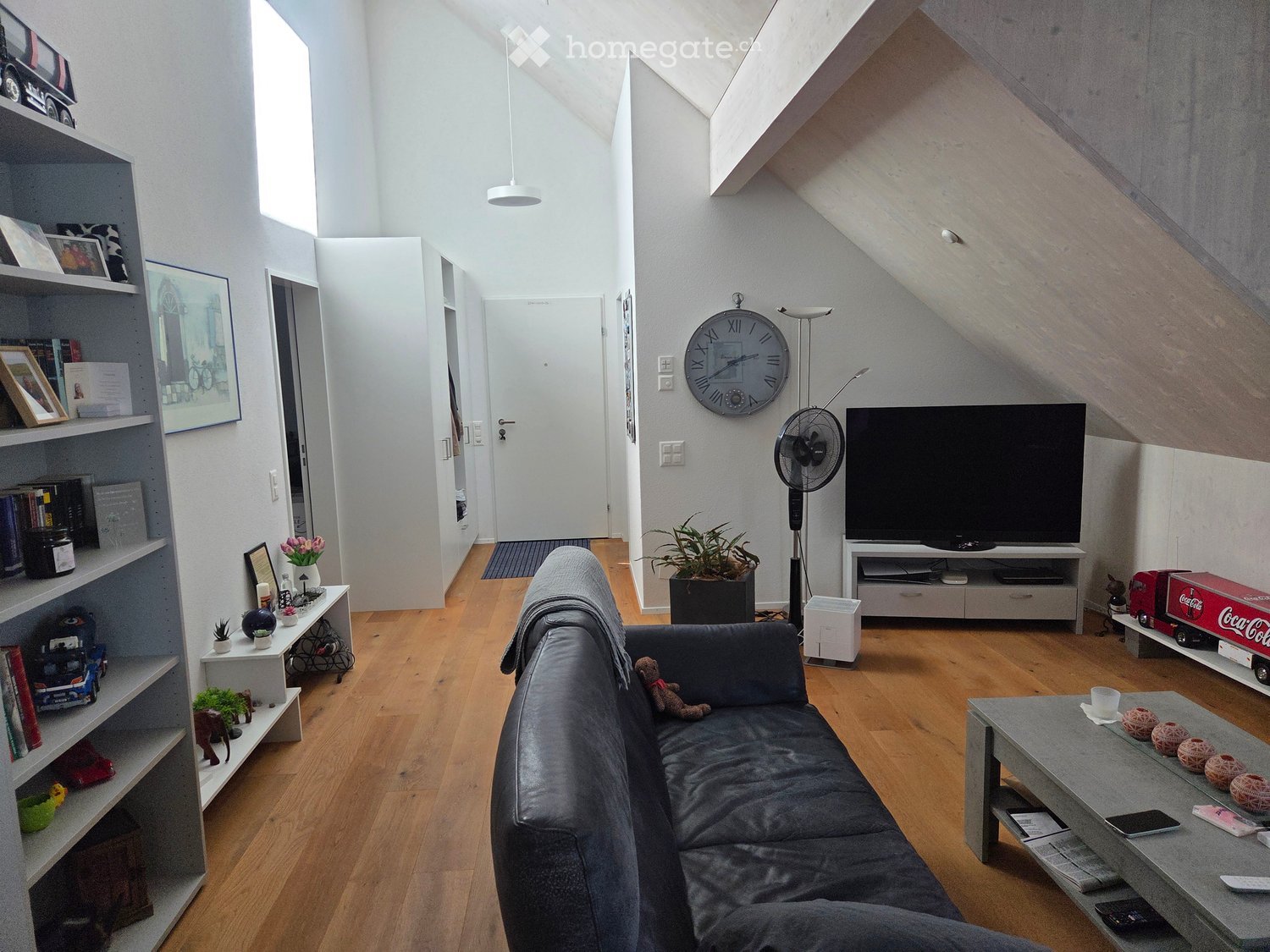 Modern living room with wooden flooring, grey sectional sofa, coffee table, TV stand, fan, clock, shelves, and decorative items.