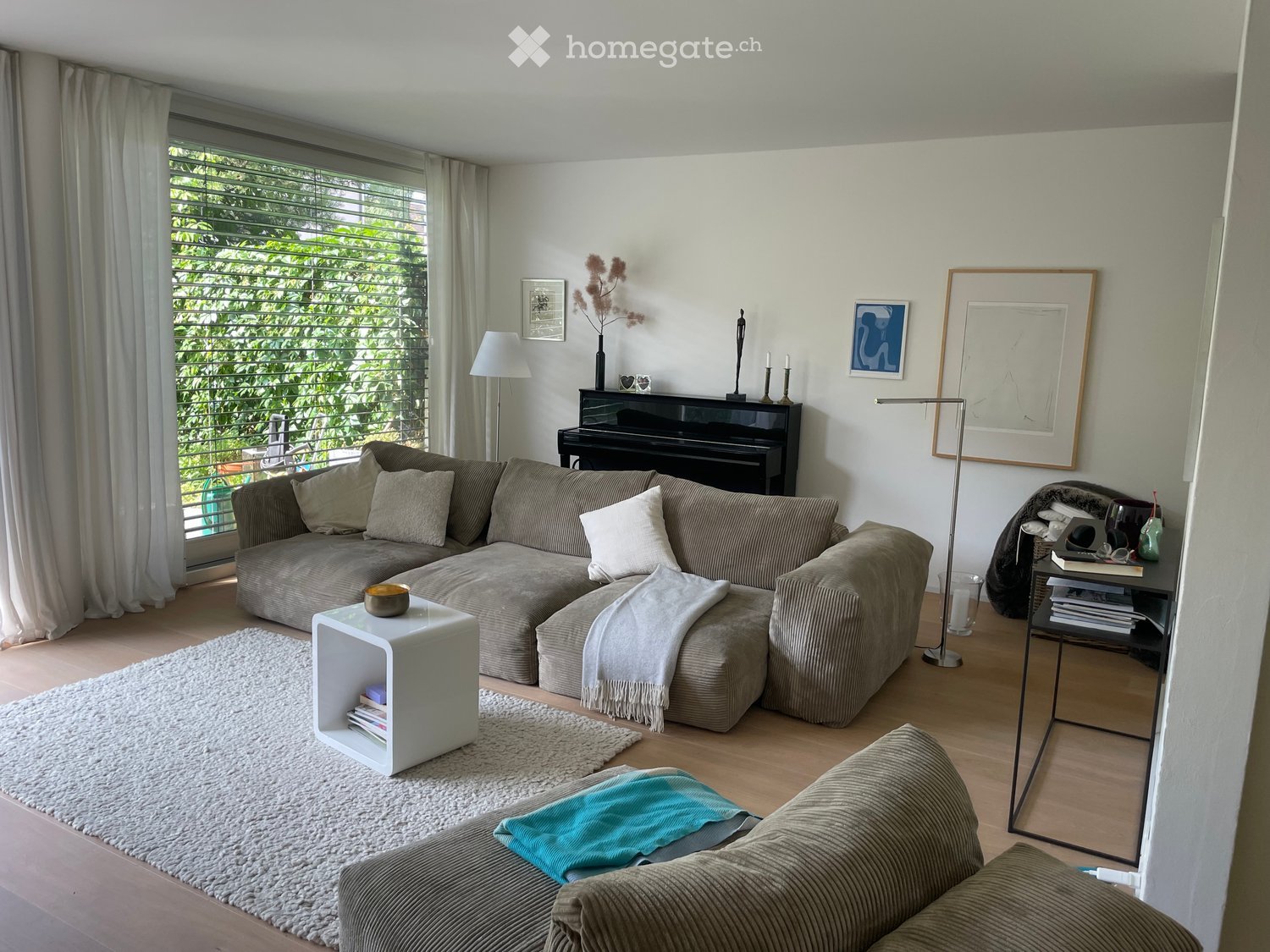 Modern living room with large windows, grey sectional sofa, white coffee table, piano, floor lamp, and decorative items.
