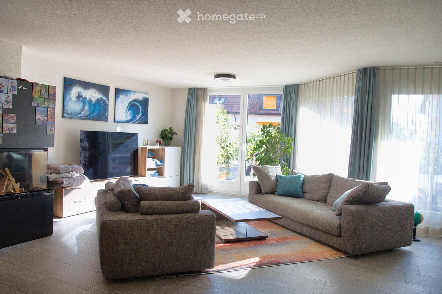 Modern living room with large windows, grey sectional sofa, coffee table, fireplace, TV, and decorative shelves.