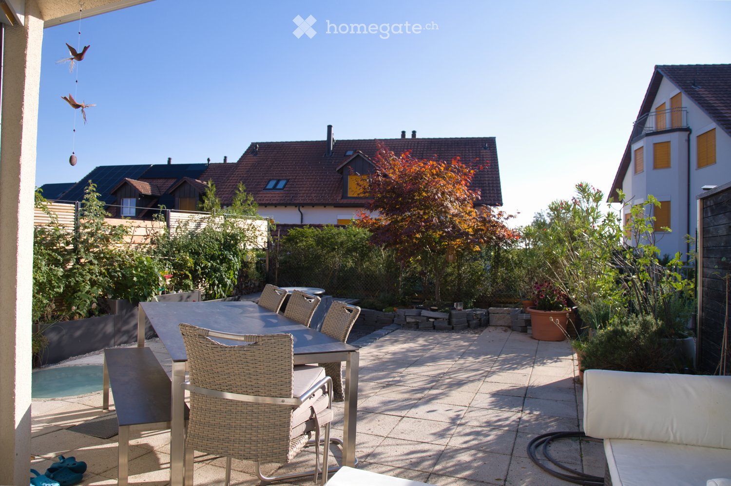 A sunny outdoor patio with a table, chairs, and a sofa, surrounded by greenery and a small swimming pool.