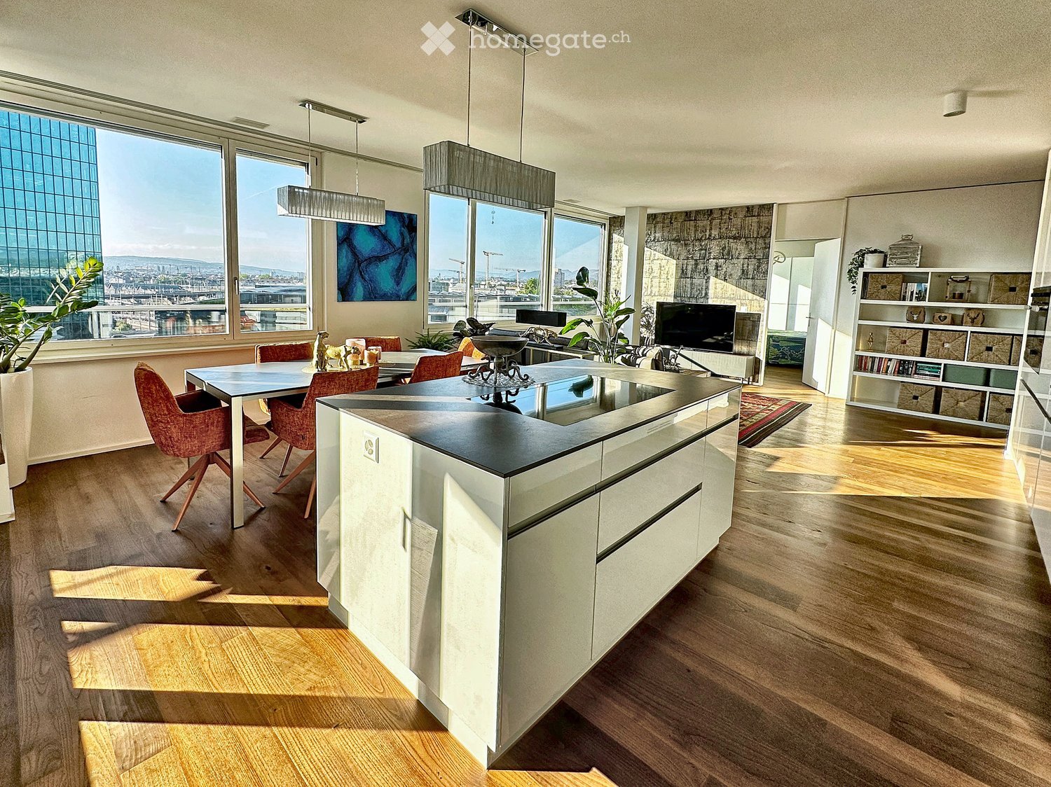 Modern kitchen with white cabinets, black countertop, pendant lights, dining area with red chairs, large windows with city view, bookshelf, and decorative plants.