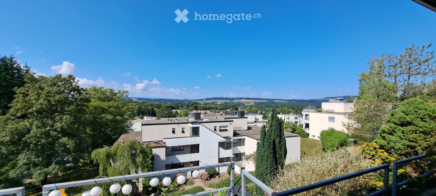 Balcony overlooking a residential area with modern apartment buildings, trees, and a clear blue sky.