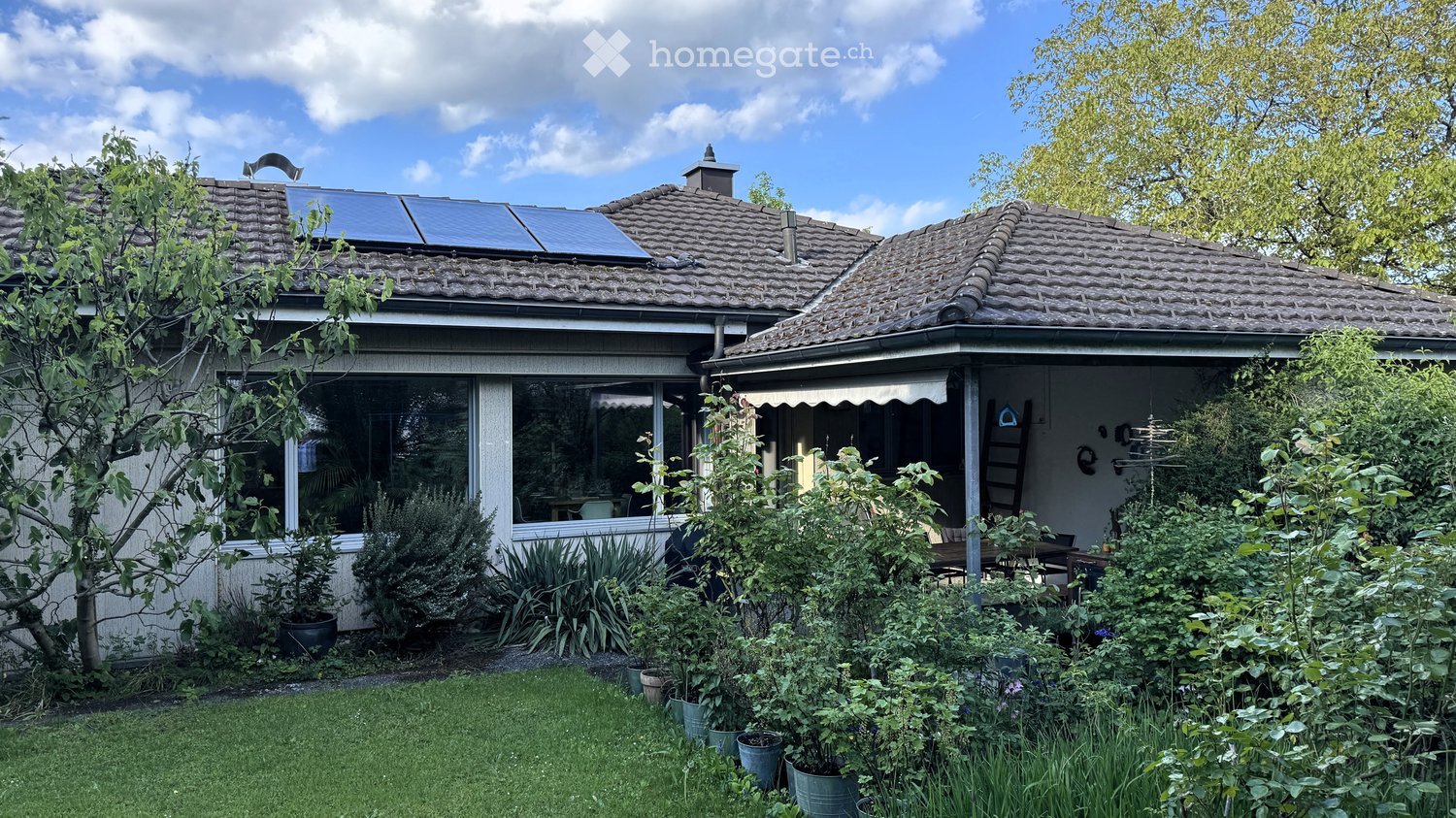 Two-story house with a tiled roof, large windows, and solar panels on the roof. The house is surrounded by a well-maintained garden with various plants and trees.