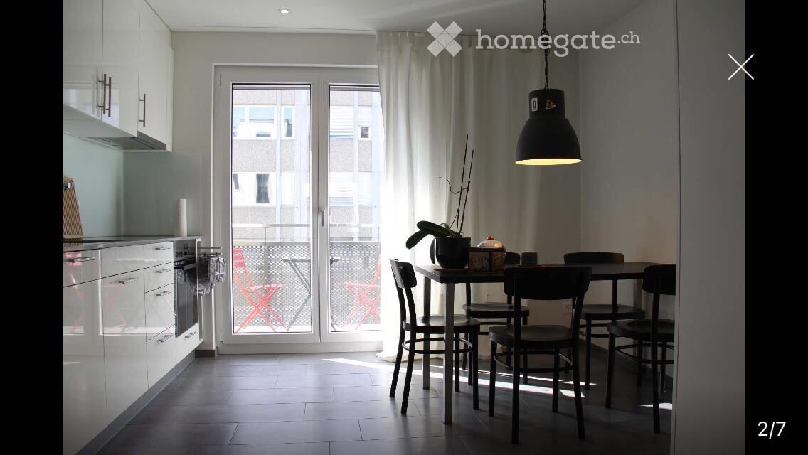 Modern kitchen and dining area with white cabinets, stainless steel appliances, a dining table with chairs, and a pendant light. Large sliding glass doors lead to a balcony with outdoor chairs.