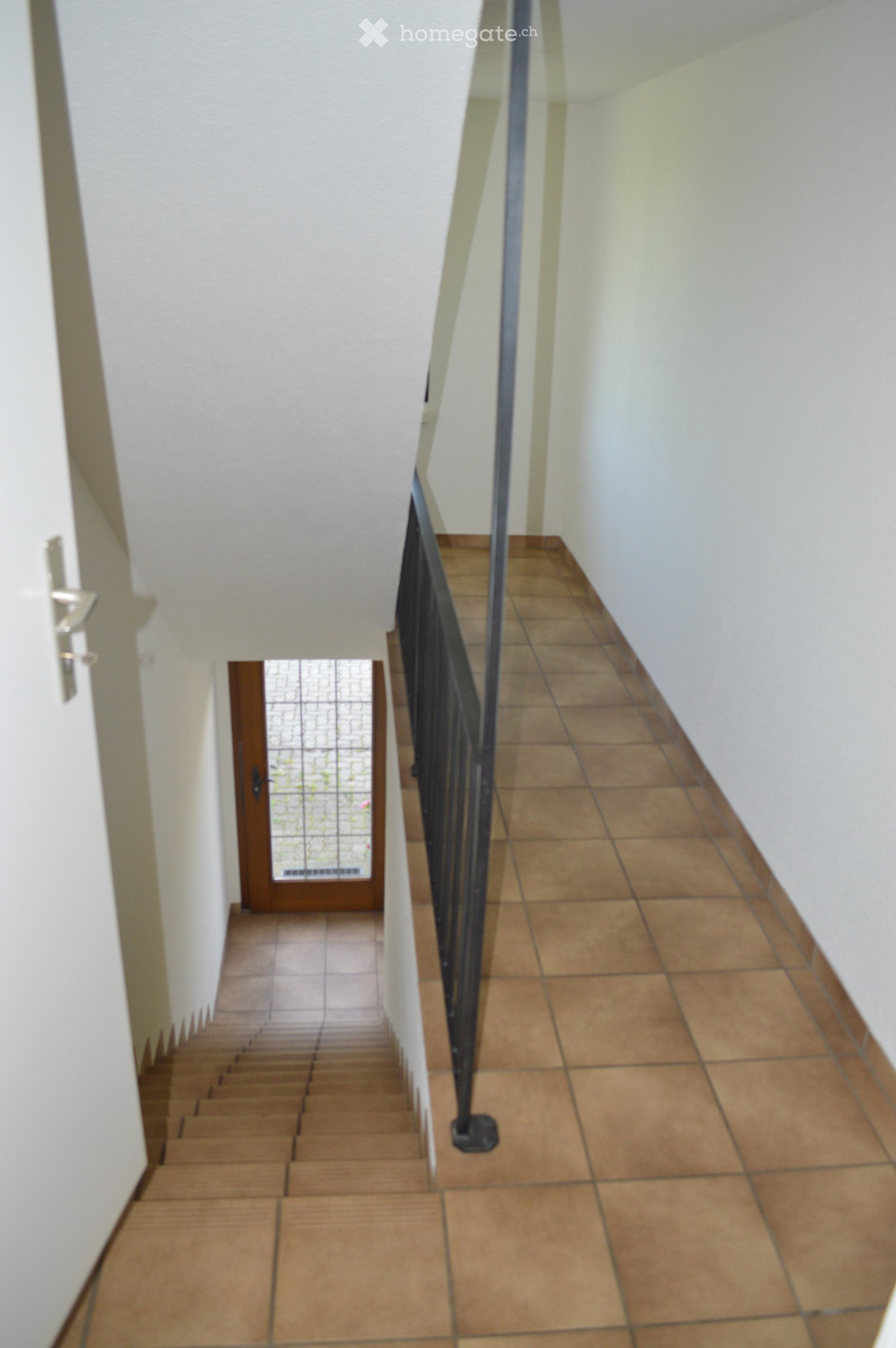 A narrow hallway with tiled flooring and white walls, featuring a staircase with a metal railing on the right side and a wooden door at the end.