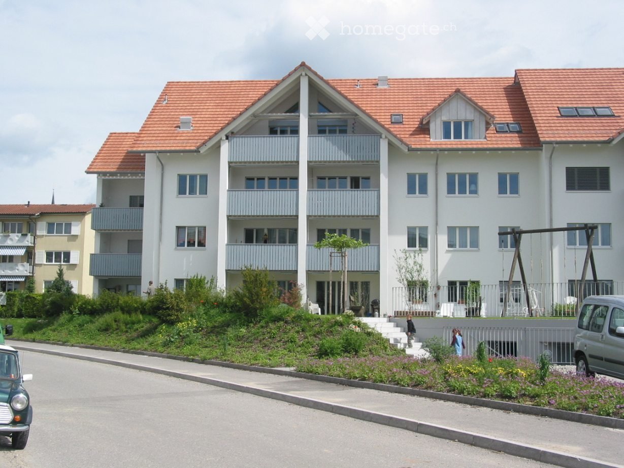 Modern multi-story apartment building with white facade, red-tiled roof, multiple balconies, and landscaped front yard with greenery and a small playground.