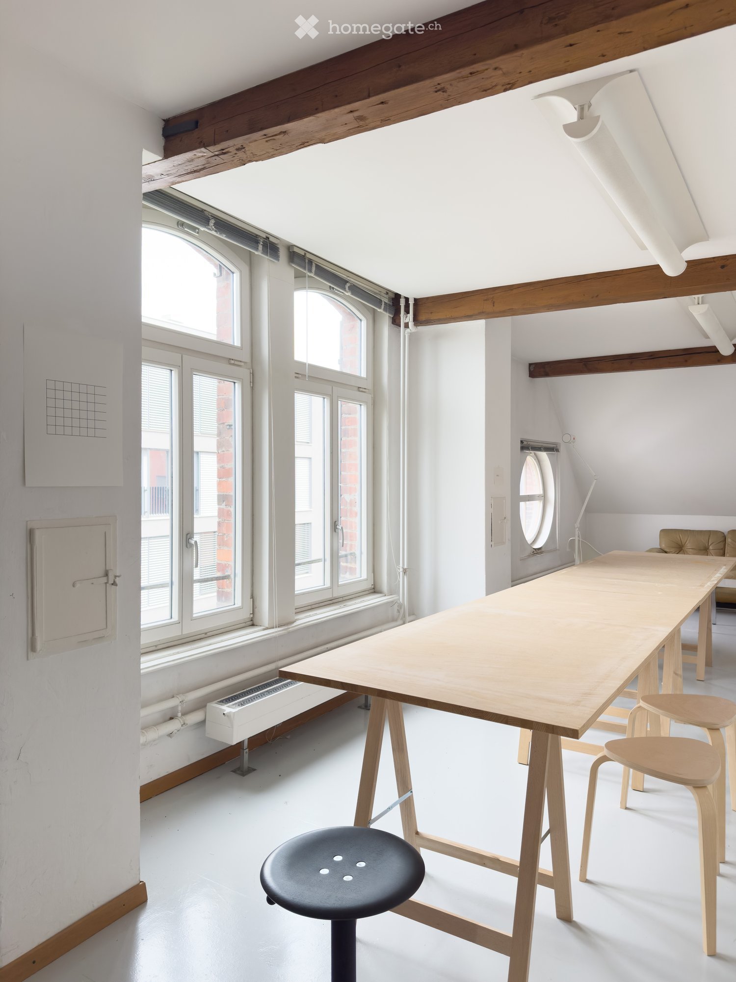 Bright, open room with exposed wooden beams, large windows, and a long wooden table with chairs and a stool.