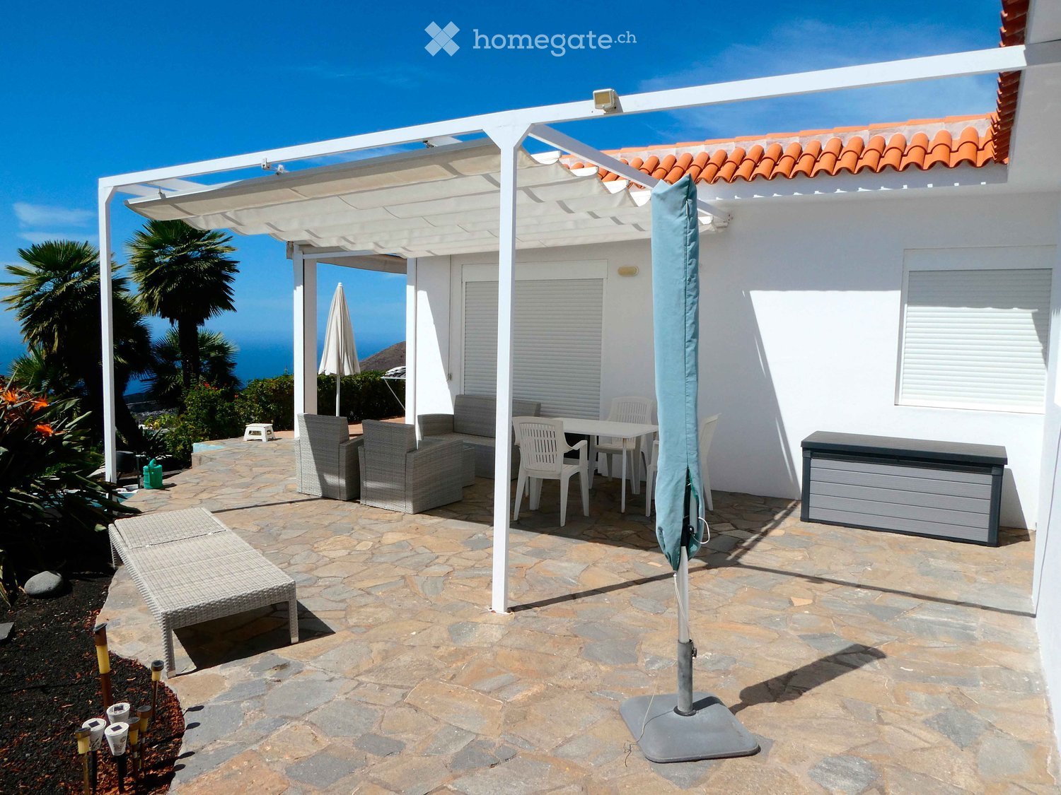 Outdoor patio with stone flooring, white pergola, sofa, table, chairs, and umbrella, overlooking the ocean.