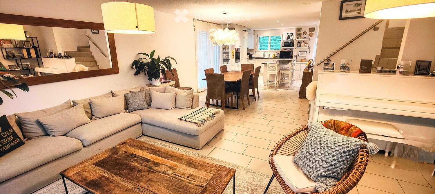 Open-plan living and dining area with a grey sectional sofa, wooden coffee table, wicker chair, dining table with chairs, and a modern kitchen with island and stools.