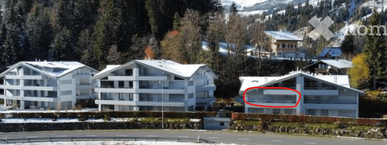 Aerial view of a multi-story residential building complex surrounded by trees and snow-covered mountains. The building has a modern design with white facades and large windows.